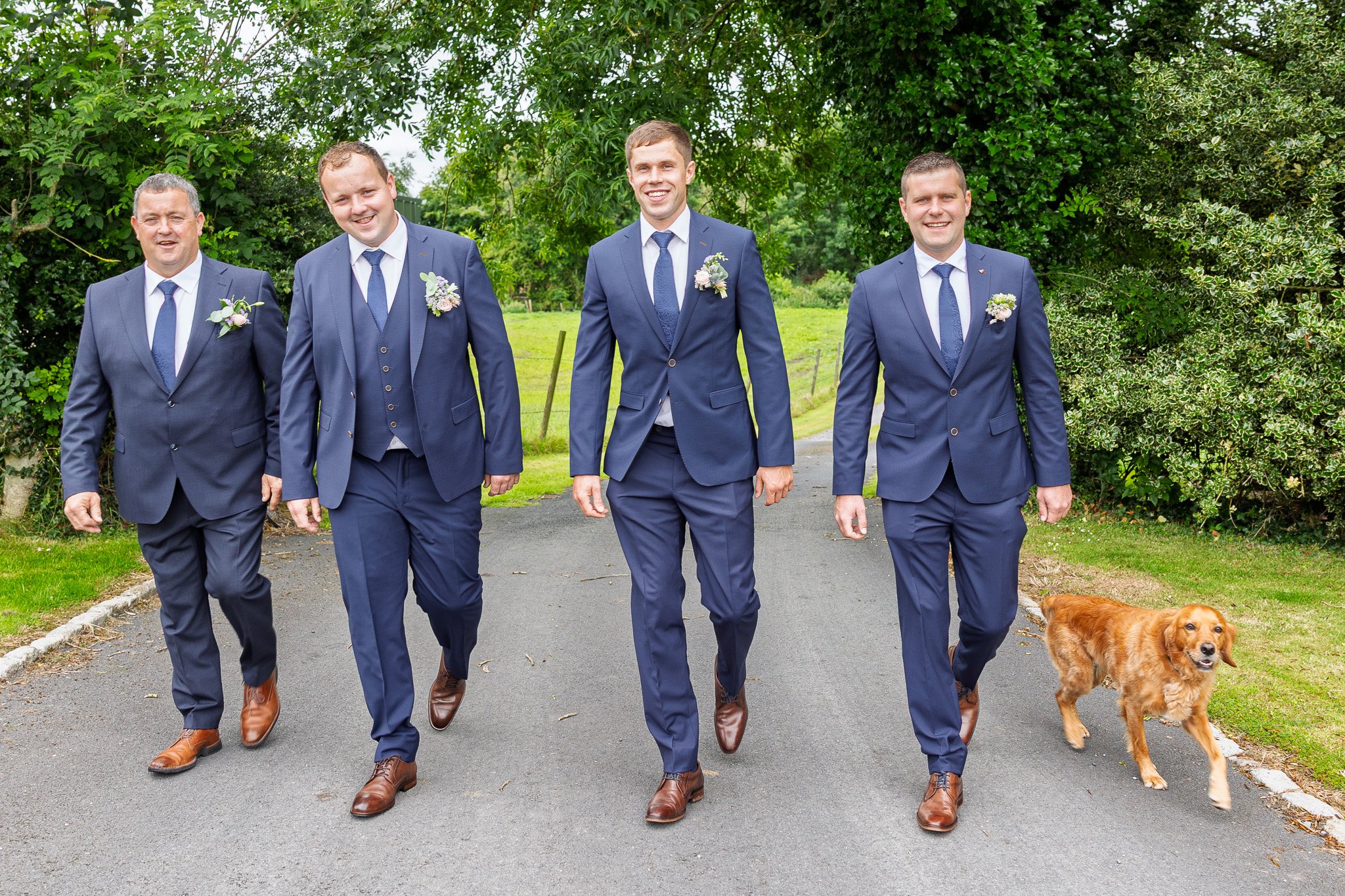 Five men in navy suits walking on a paved path outdoors, with a dog on the right. Green trees and grass are in the background.
