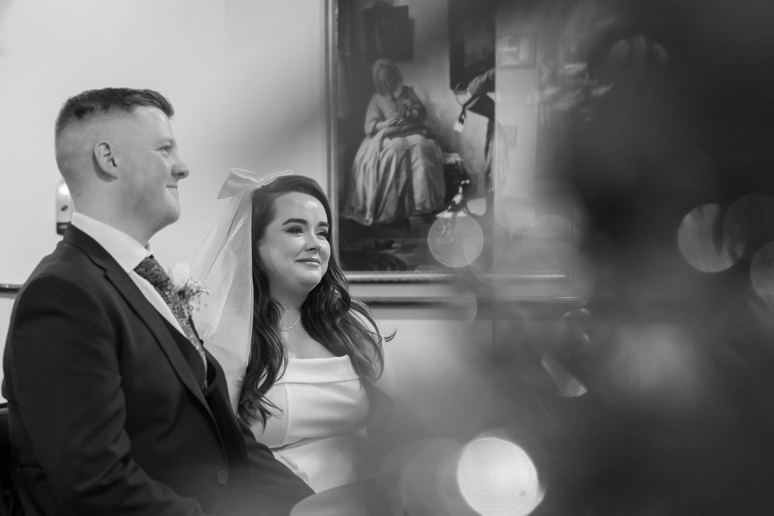 A bride and groom sitting together during their wedding ceremony. The groom is smiling, and the bride appears emotional. A painting of a woman in a dress hangs on the wall behind them.