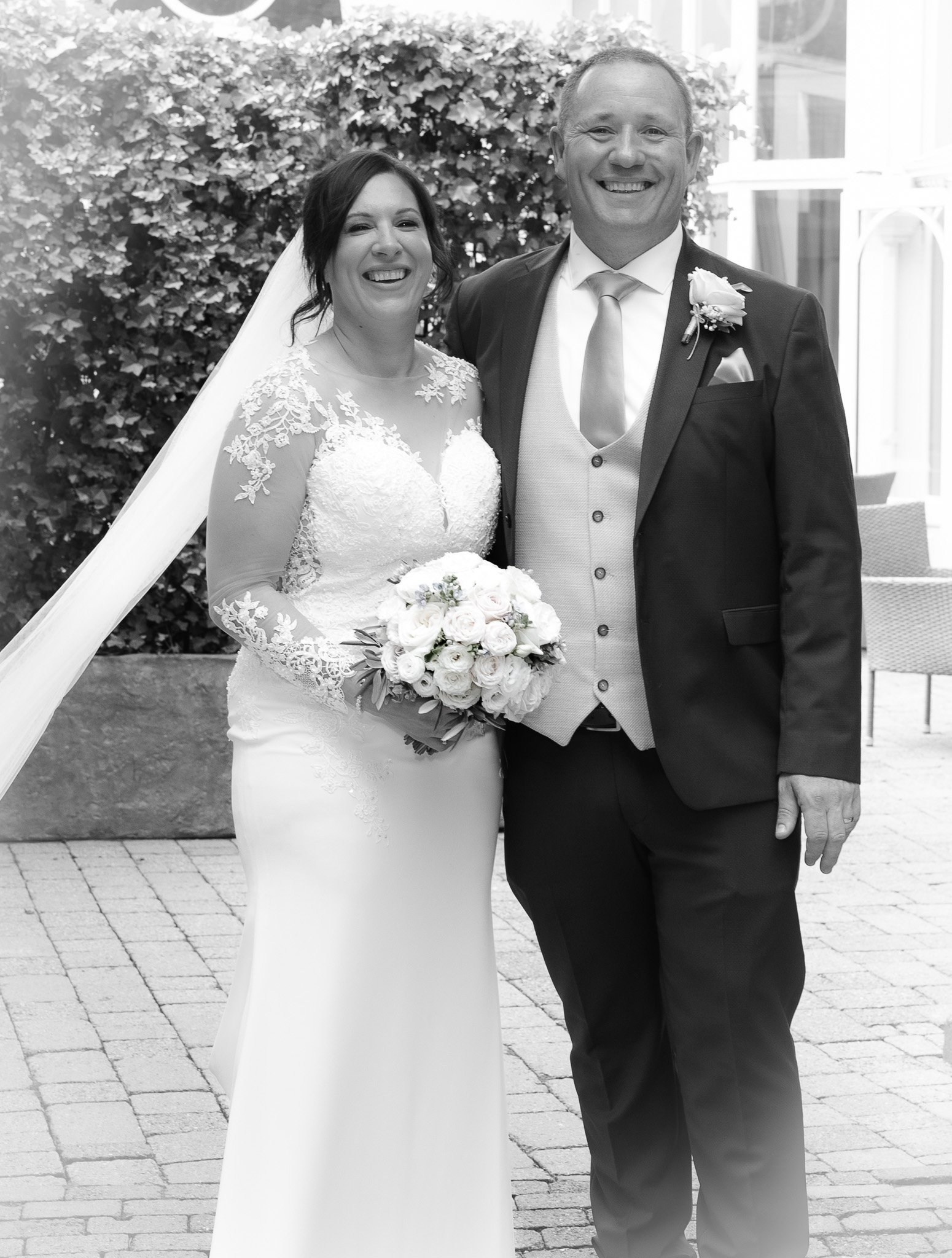 Black and white photo of a happy bride in a lace wedding gown holding a bouquet and a smiling groom in a suit with a boutonniere, standing outdoors.