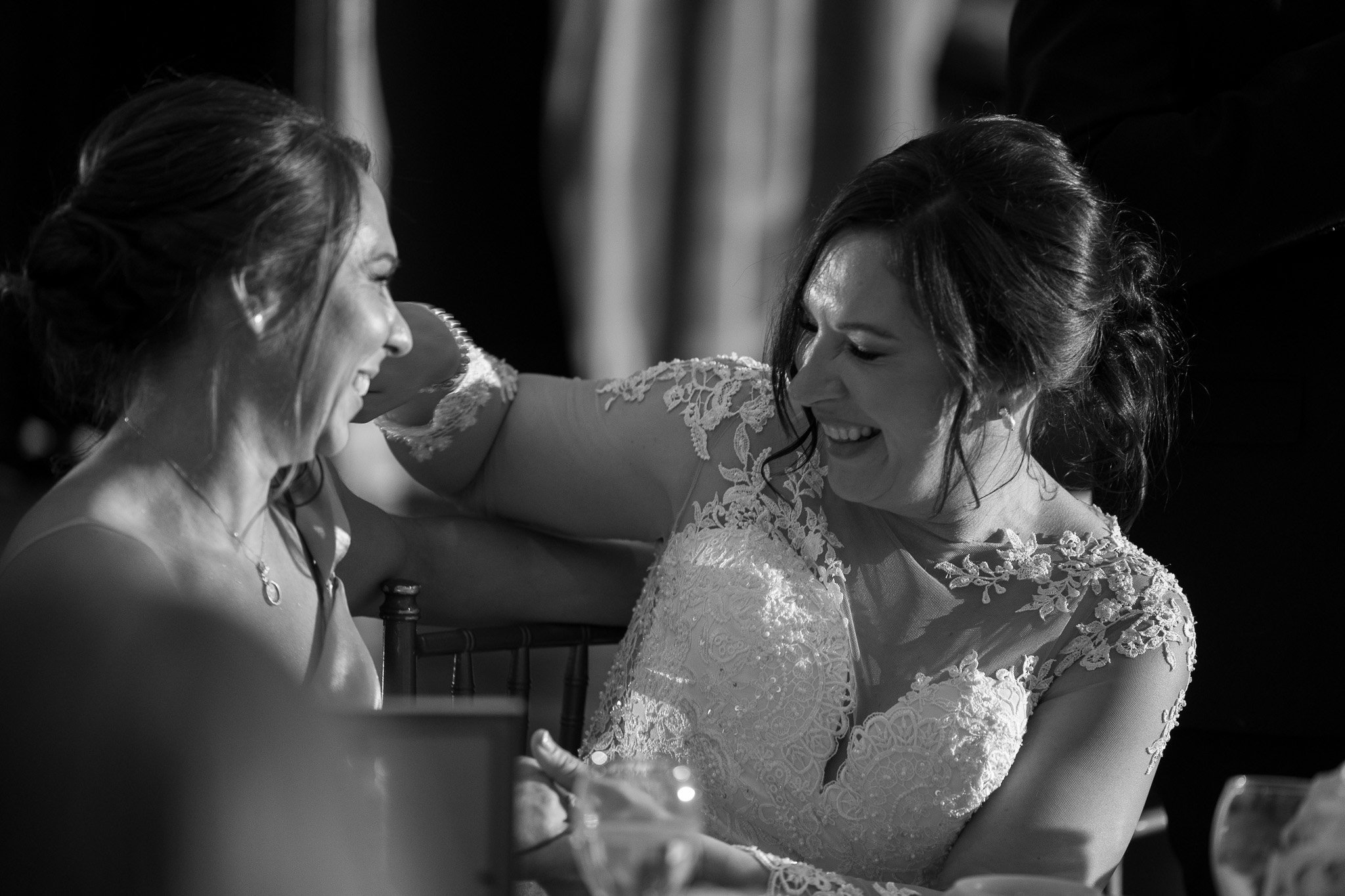 Two women in wedding dresses smiling and laughing, sitting closely together at a celebration or wedding reception.