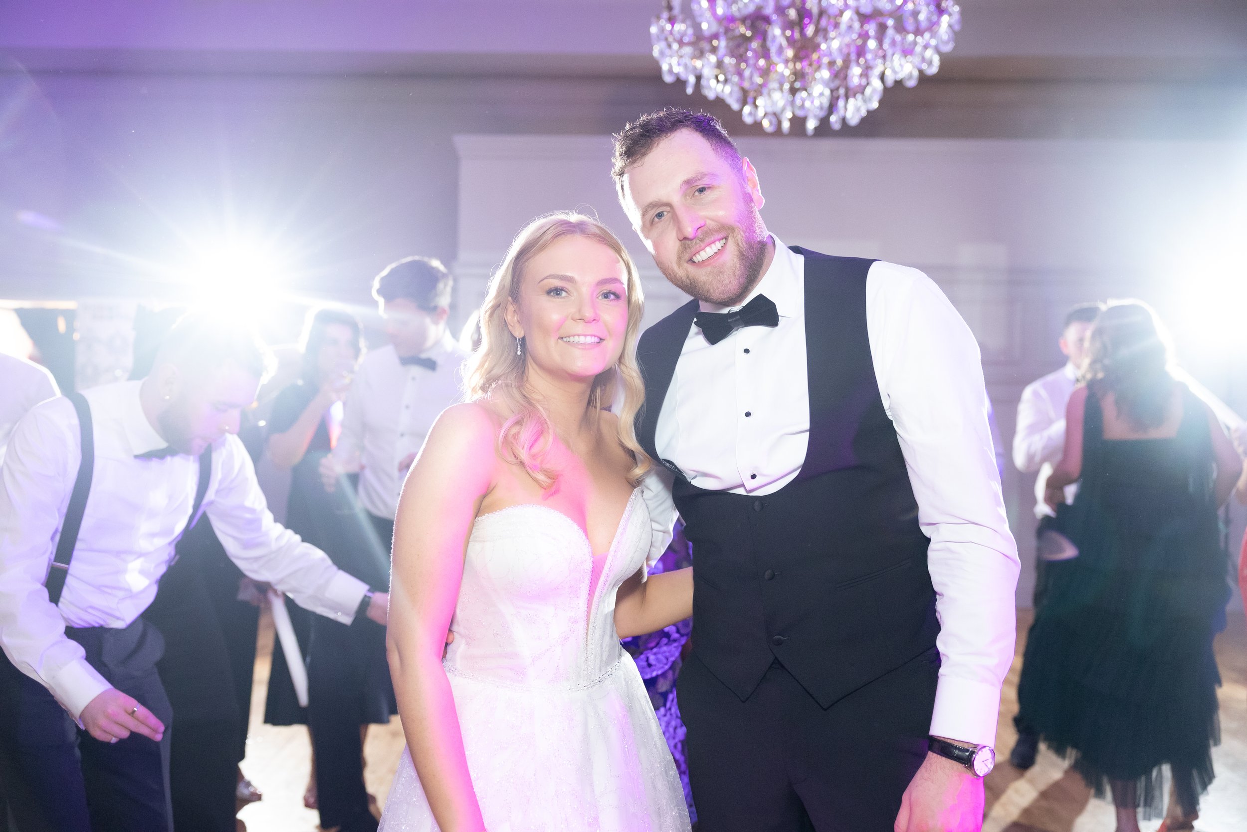 A smiling bride and groom in wedding attire at a reception, dancing and celebrating with friends in the background, illuminated by bright lights and a chandelier.