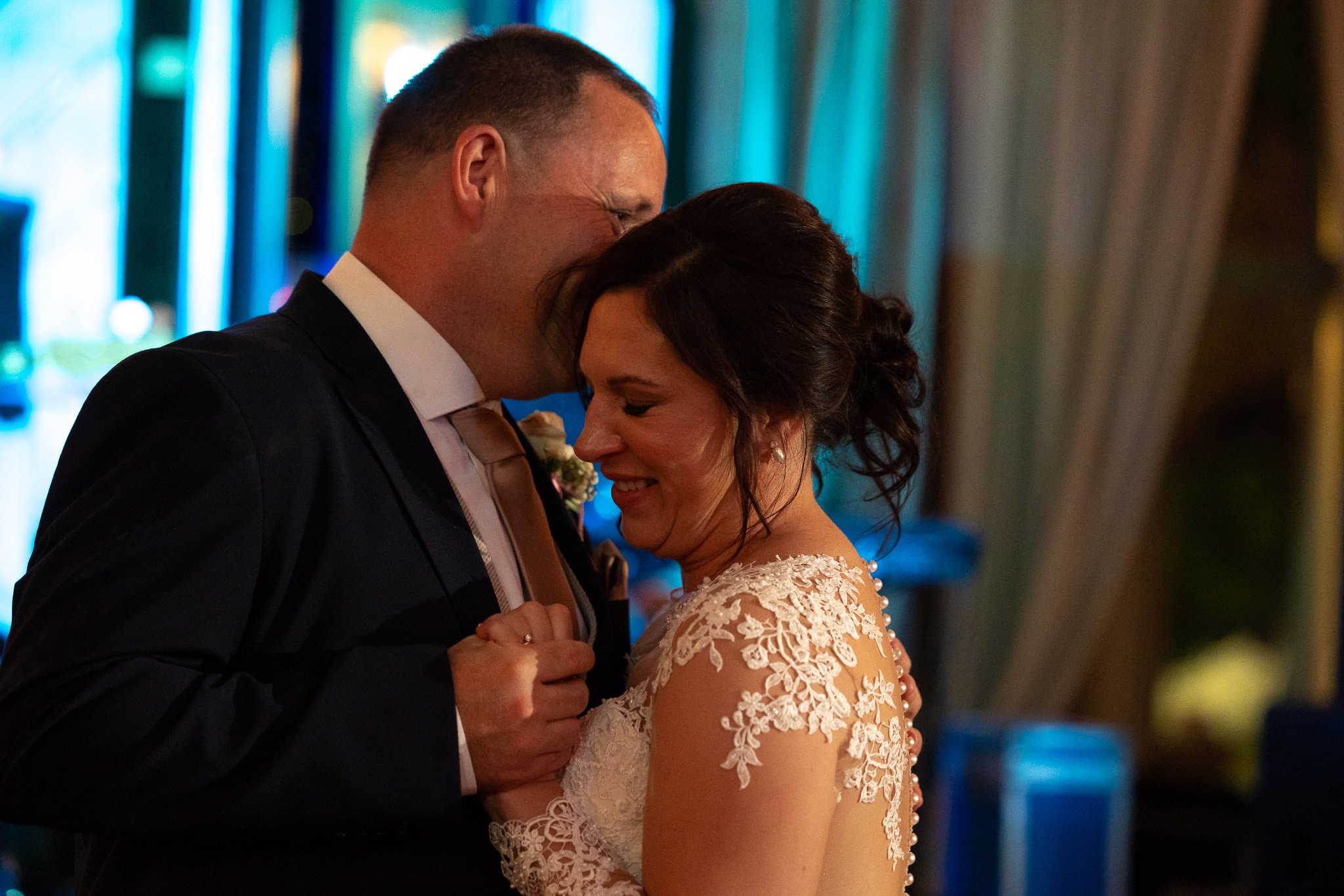 A couple dancing closely at their wedding, with the groom whispering into the bride's ear, both smiling with joy.