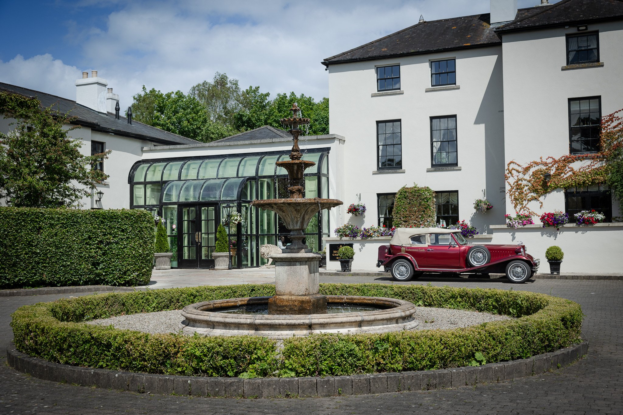 A fountain with water bubbling at the center in front of a well-maintained garden with a vintage red car parked beside a white building with black-trimmed windows and hanging flower pots.