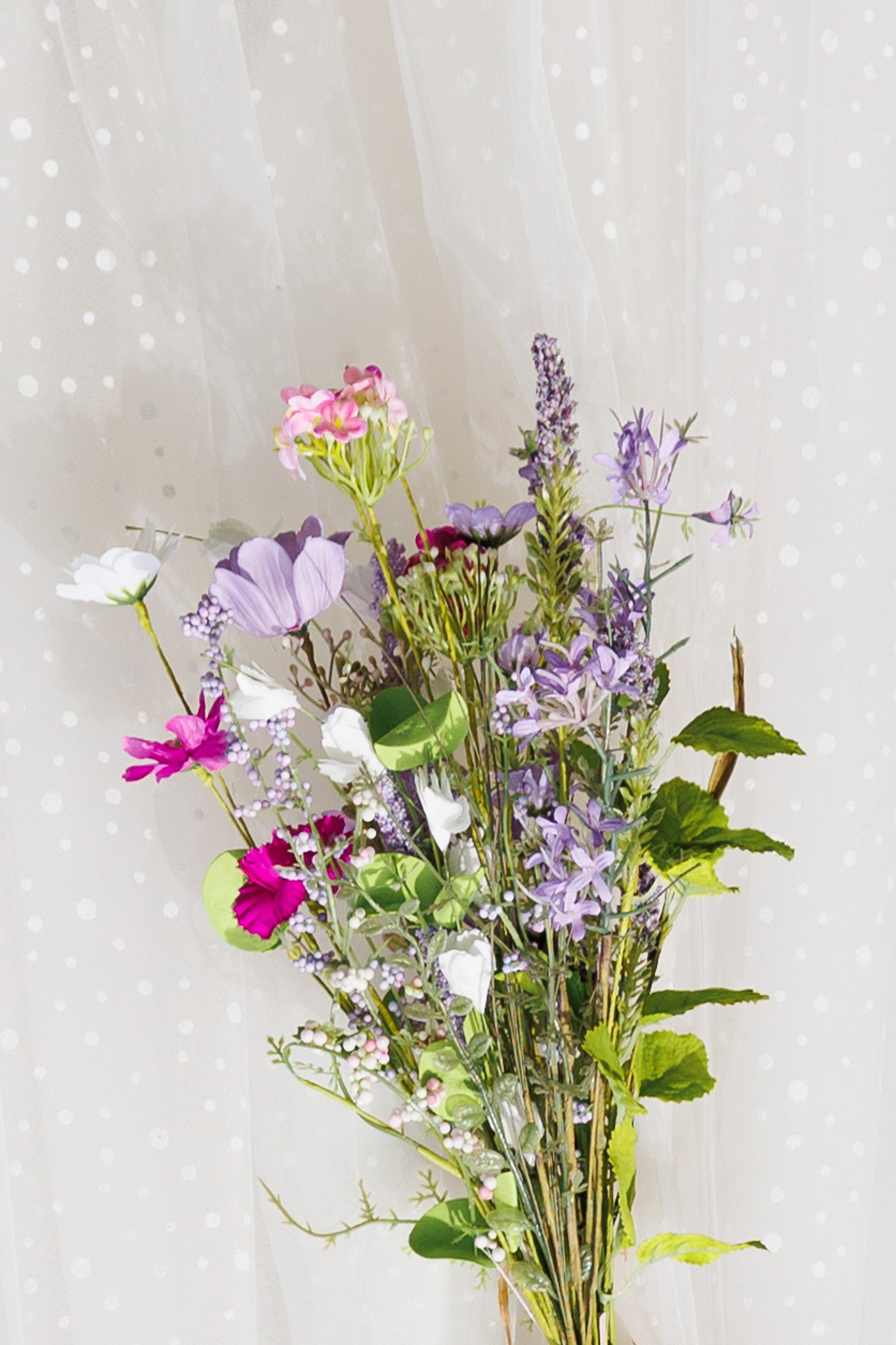 A bouquet of mixed flowers in pink, purple, white, and green, with a beige dotted background.