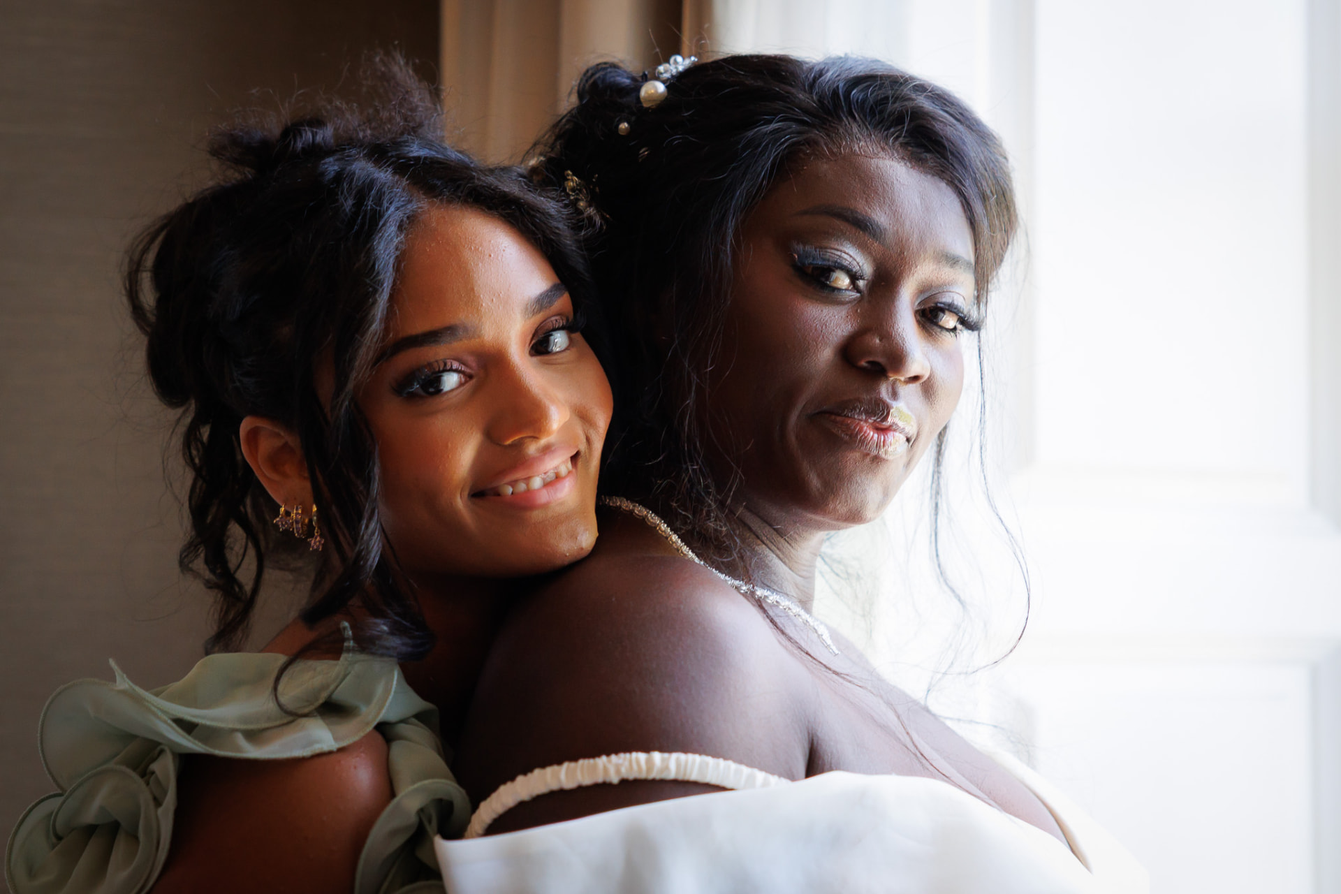 Bride and bridesmaid cheek to cheek in soft window light