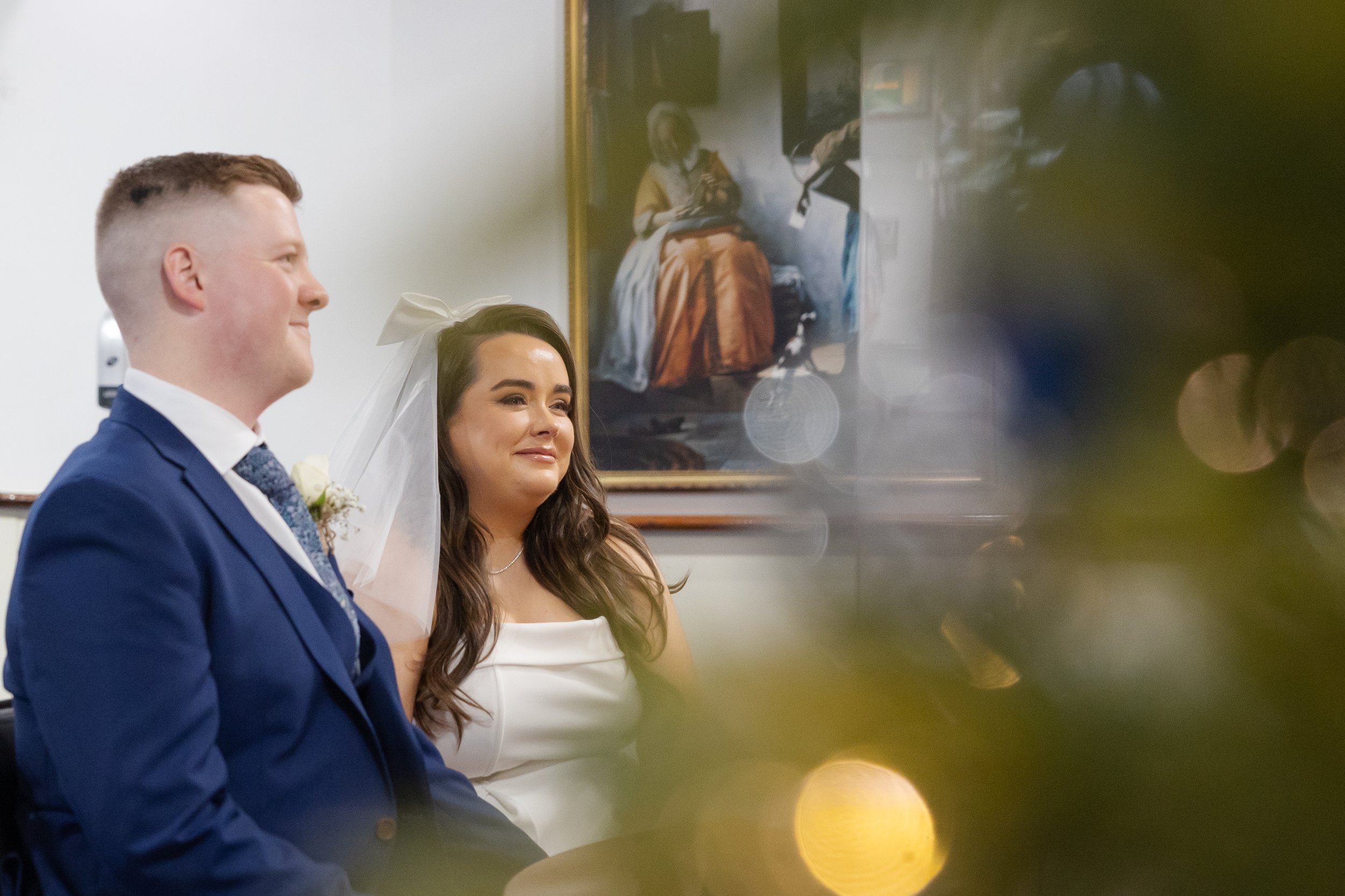 A groom in a blue suit and a bride with long dark hair and a white dress, sitting together during their wedding ceremony, with emotional expressions.