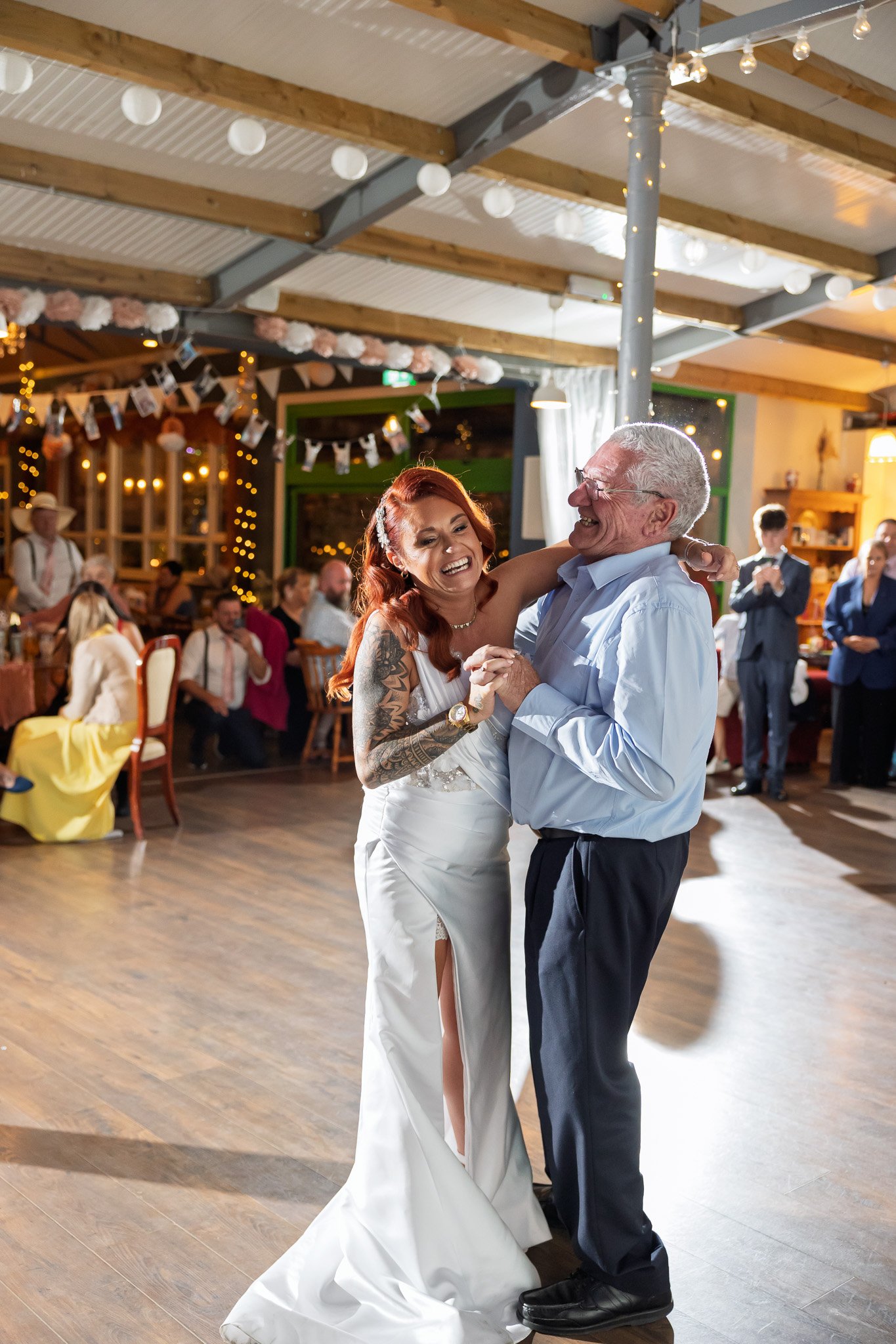 A bride and an older man are dancing and smiling at a wedding reception in a decorated hall with candles and string lights.