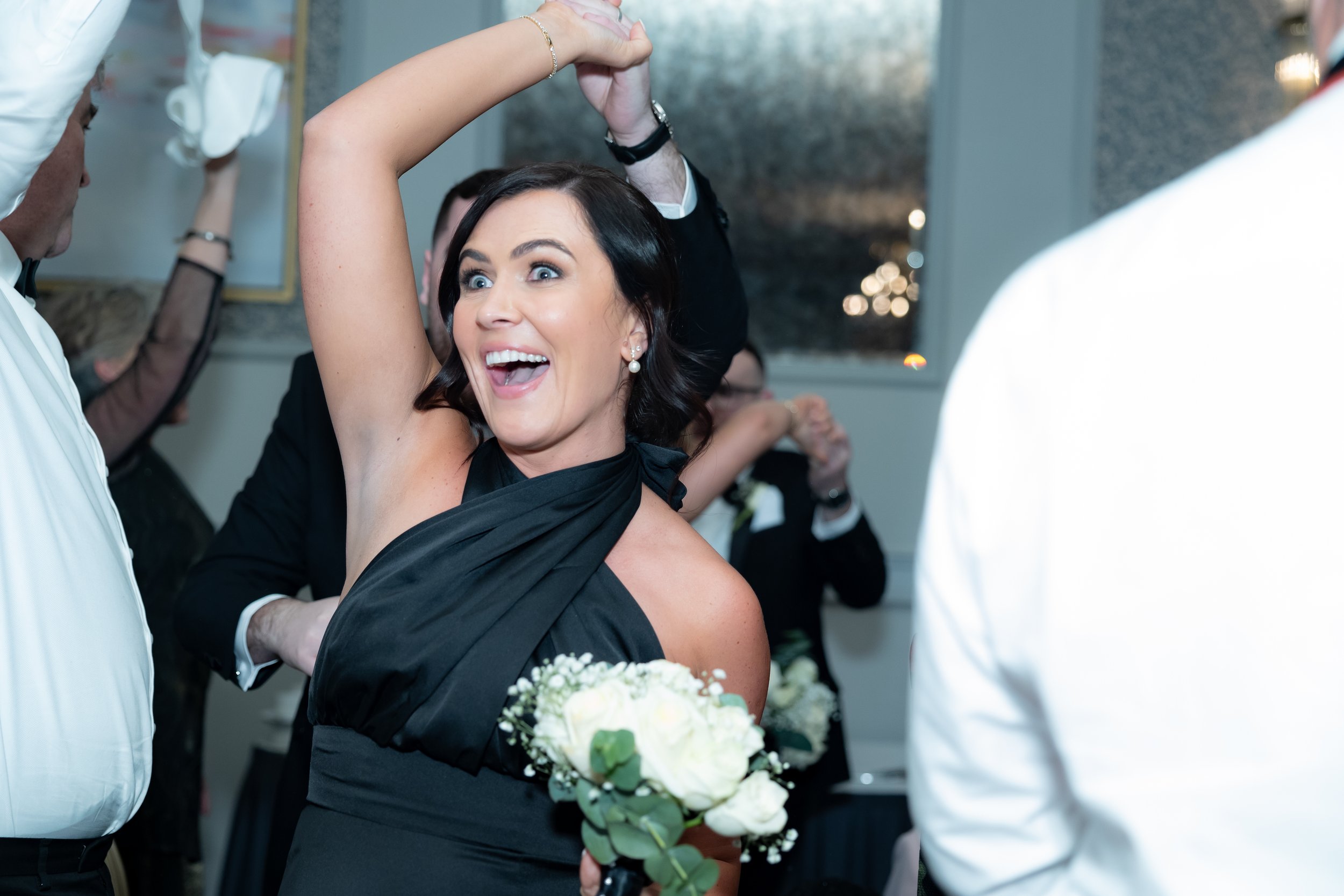 A woman in a black dress smiling and holding a bouquet of white flowers, dancing or celebrating with people at a social event.