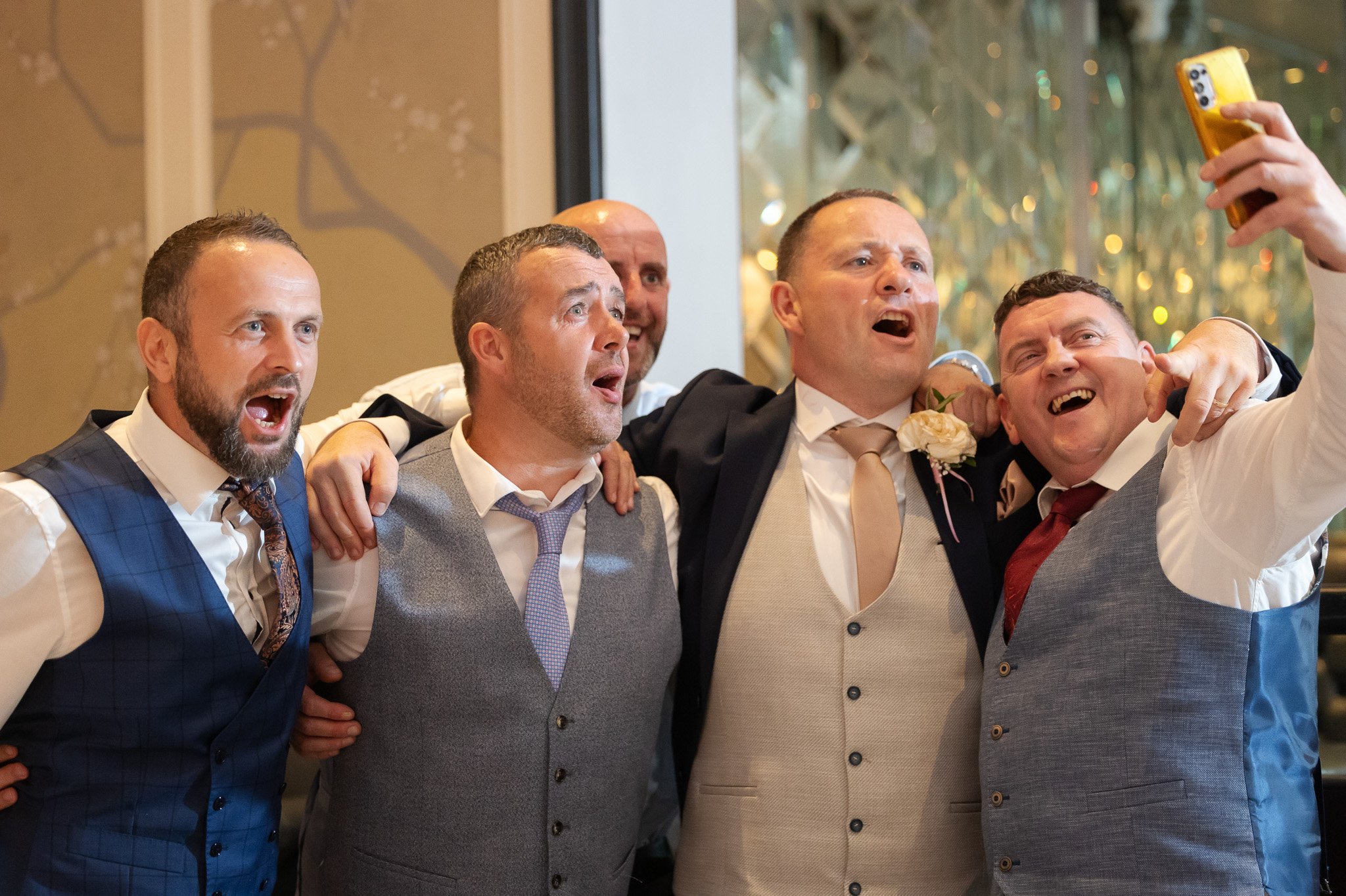 Five men dressed in formal wedding attire taking a selfie together, celebrating, smiling, with a background featuring decorative lights and foliage.