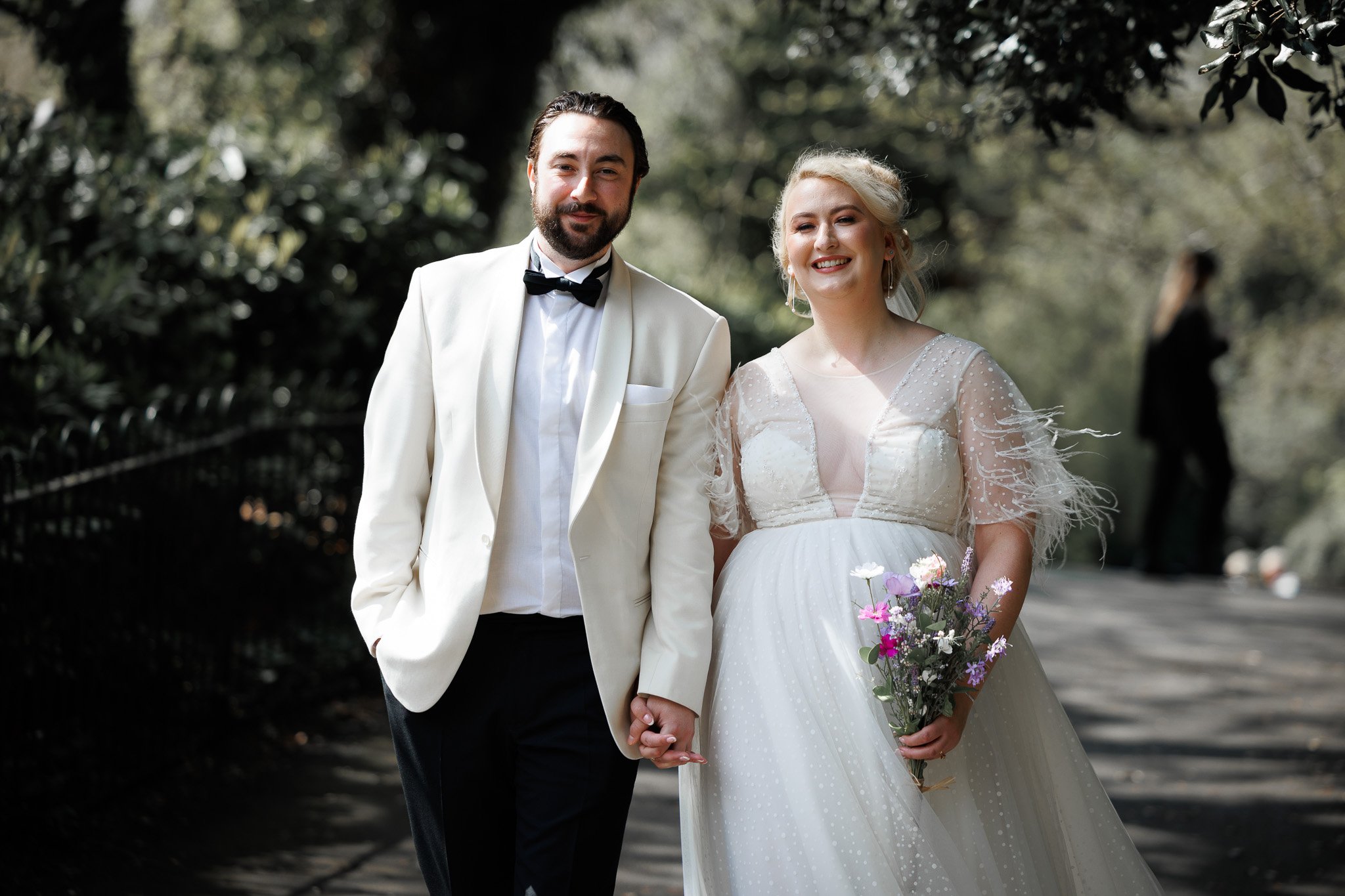 A newlywed couple walking hand in hand outdoors on a sunny day, the bride holding a bouquet of flowers, both smiling.