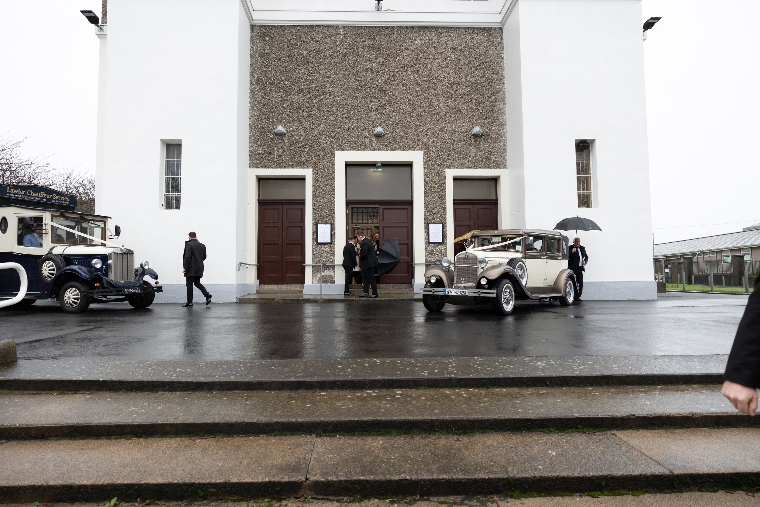 People dressed in formal attire standing outside a white church or chapel on a rainy day, with vintage cars parked in front.