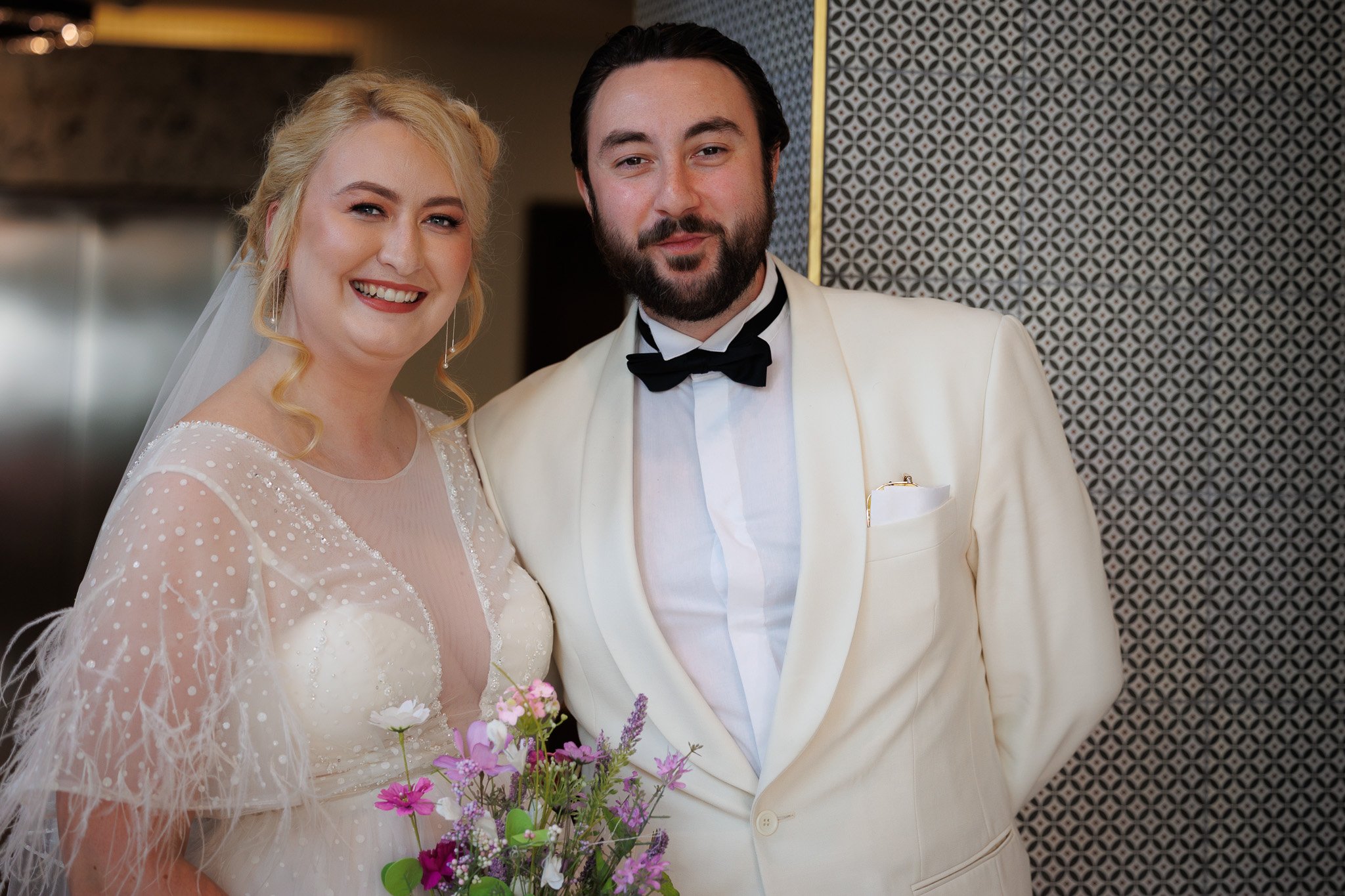 Happy bride in a white wedding dress holding a bouquet of flowers with groom in a white tuxedo and black bow tie standing close together.