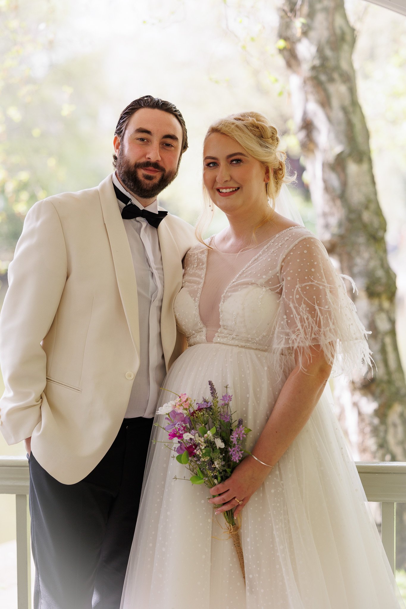 A bride and groom standing together outdoors, smiling, with the bride holding a bouquet of flowers. The groom is wearing a cream tuxedo jacket with a black bow tie, and the bride is wearing a white wedding dress with lace and tulle details.