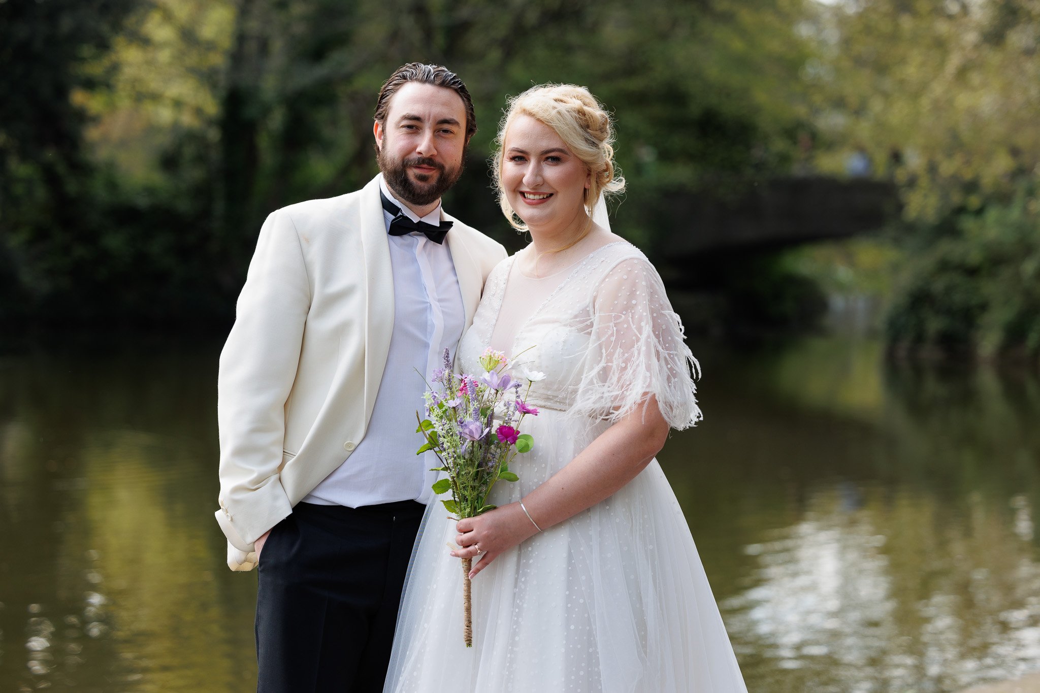 A newlywed couple in wedding attire outdoors, standing by a body of water with trees in the background. The woman is holding a bouquet of flowers and smiling, while the man is dressed in a tuxedo and smiling.