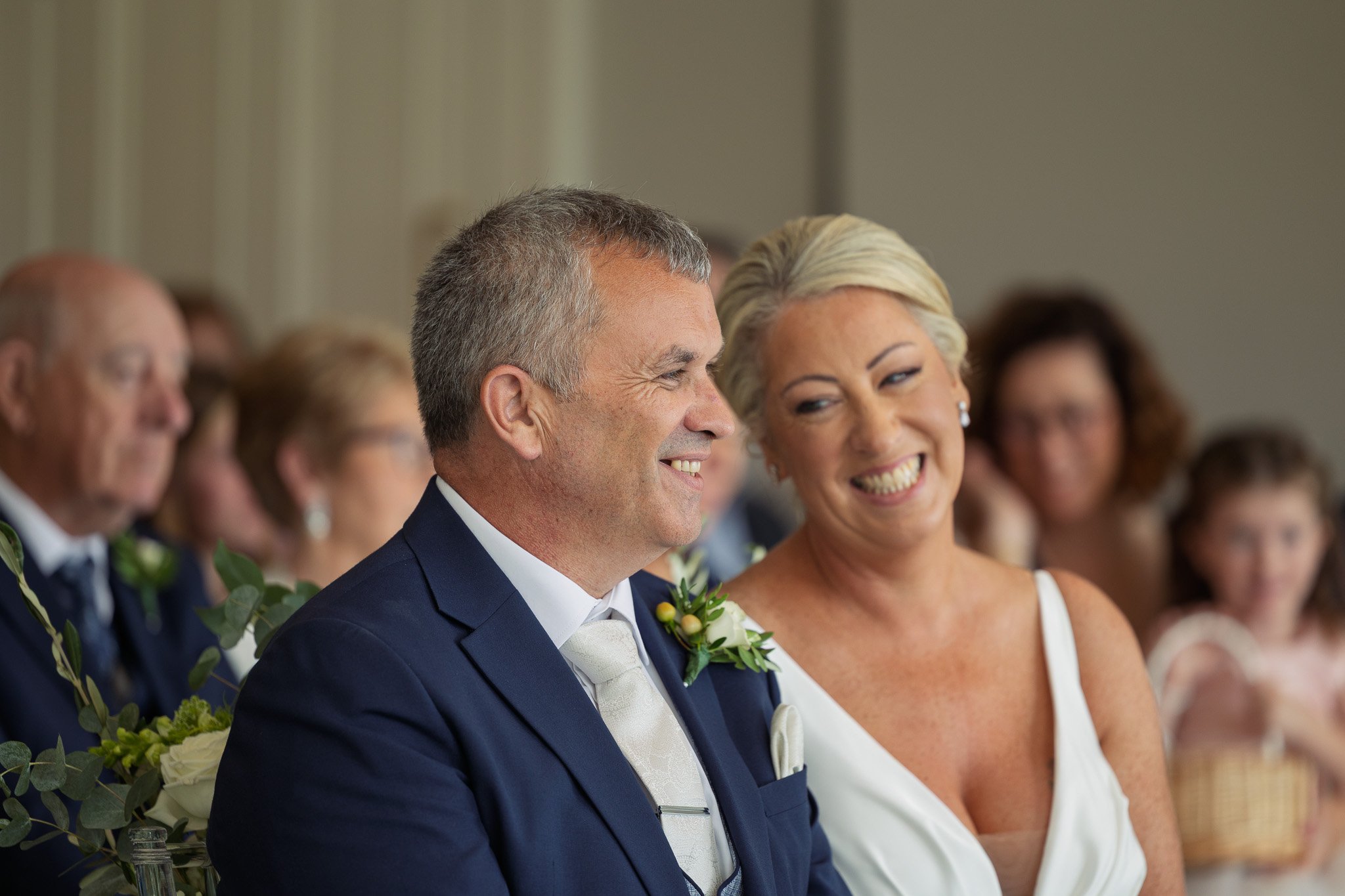 A smiling middle-aged man in a navy suit with a white tie and a boutonniere, and a smiling middle-aged woman with short blonde hair in a white dress, sitting together at a wedding ceremony, surrounded by seated guests.