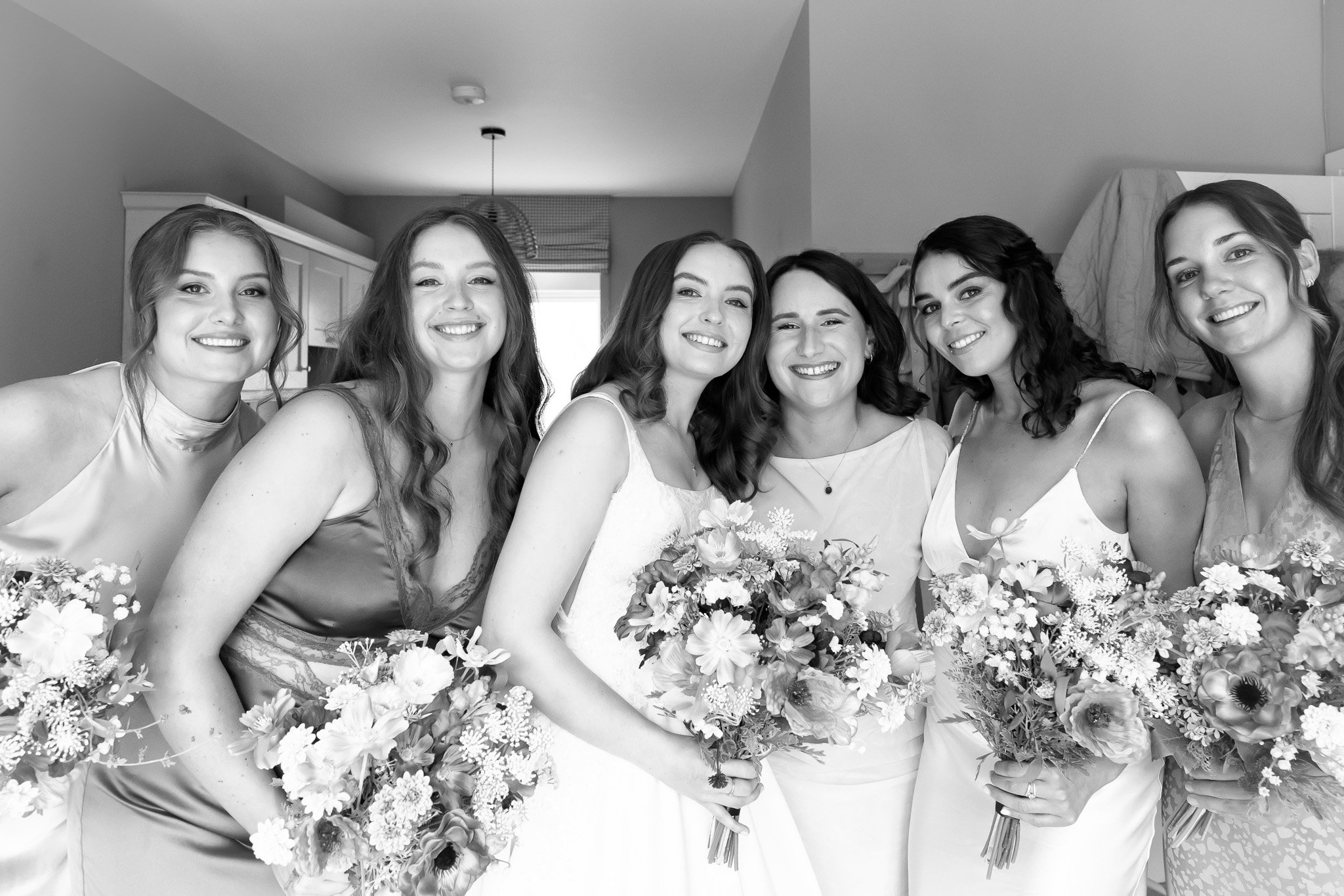 Black and white photo of six smiling women at a wedding, holding bouquets of flowers, standing closely together indoors.