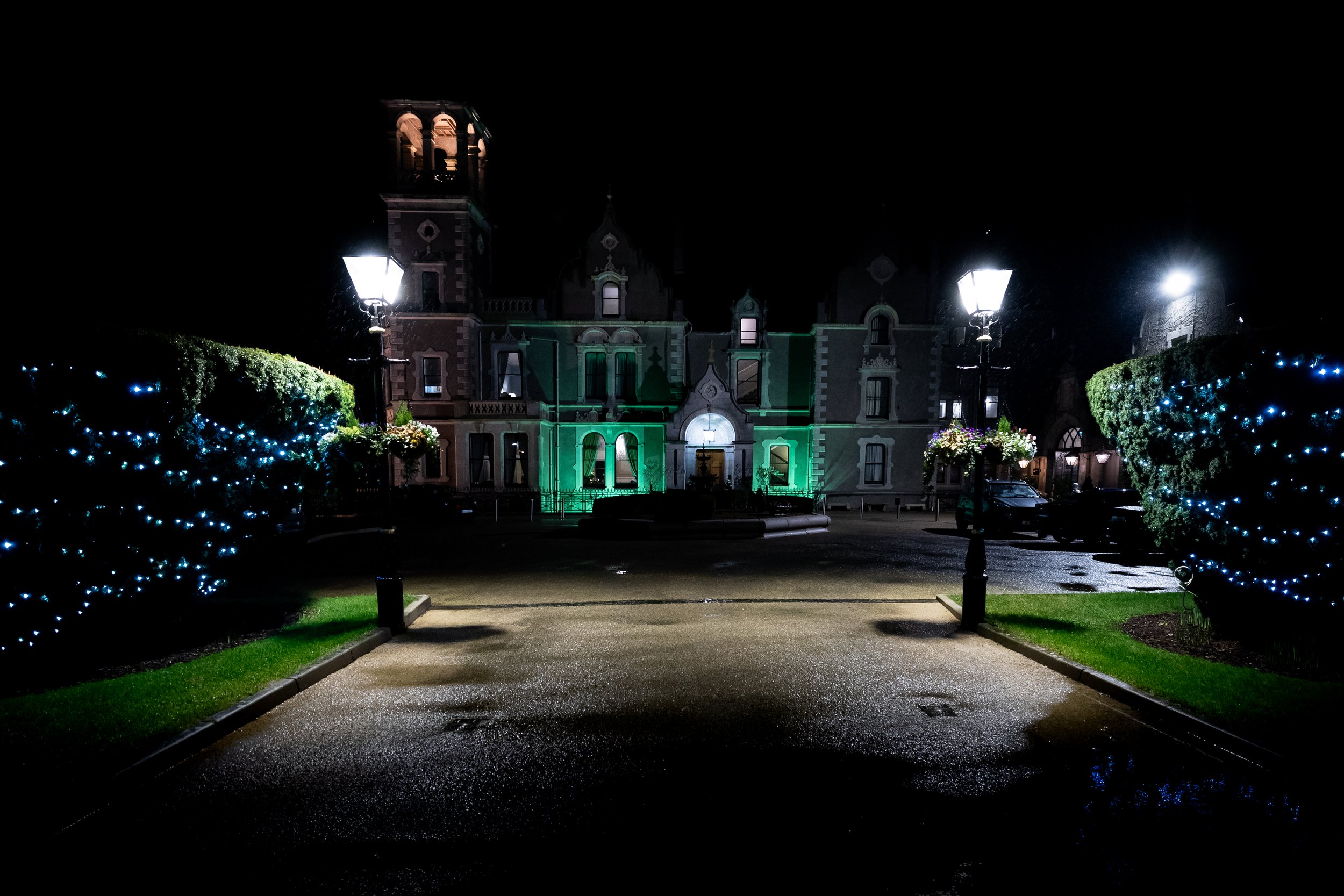 Night view of a grand, historic building illuminated with green and white lights, flanked by two lamp posts, with decorated bushes on either side.