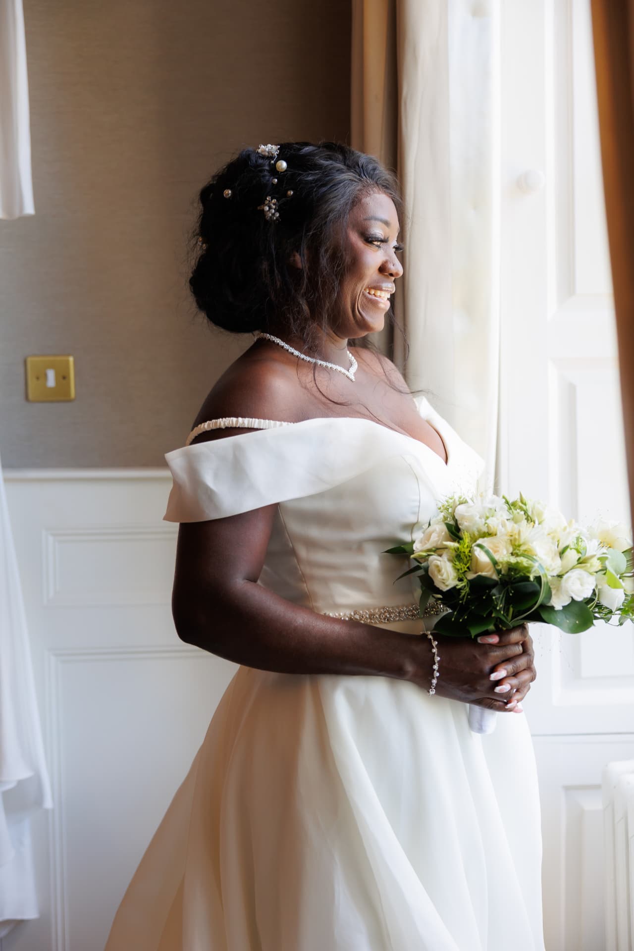 Bride laughing by a window holding a white bouquet