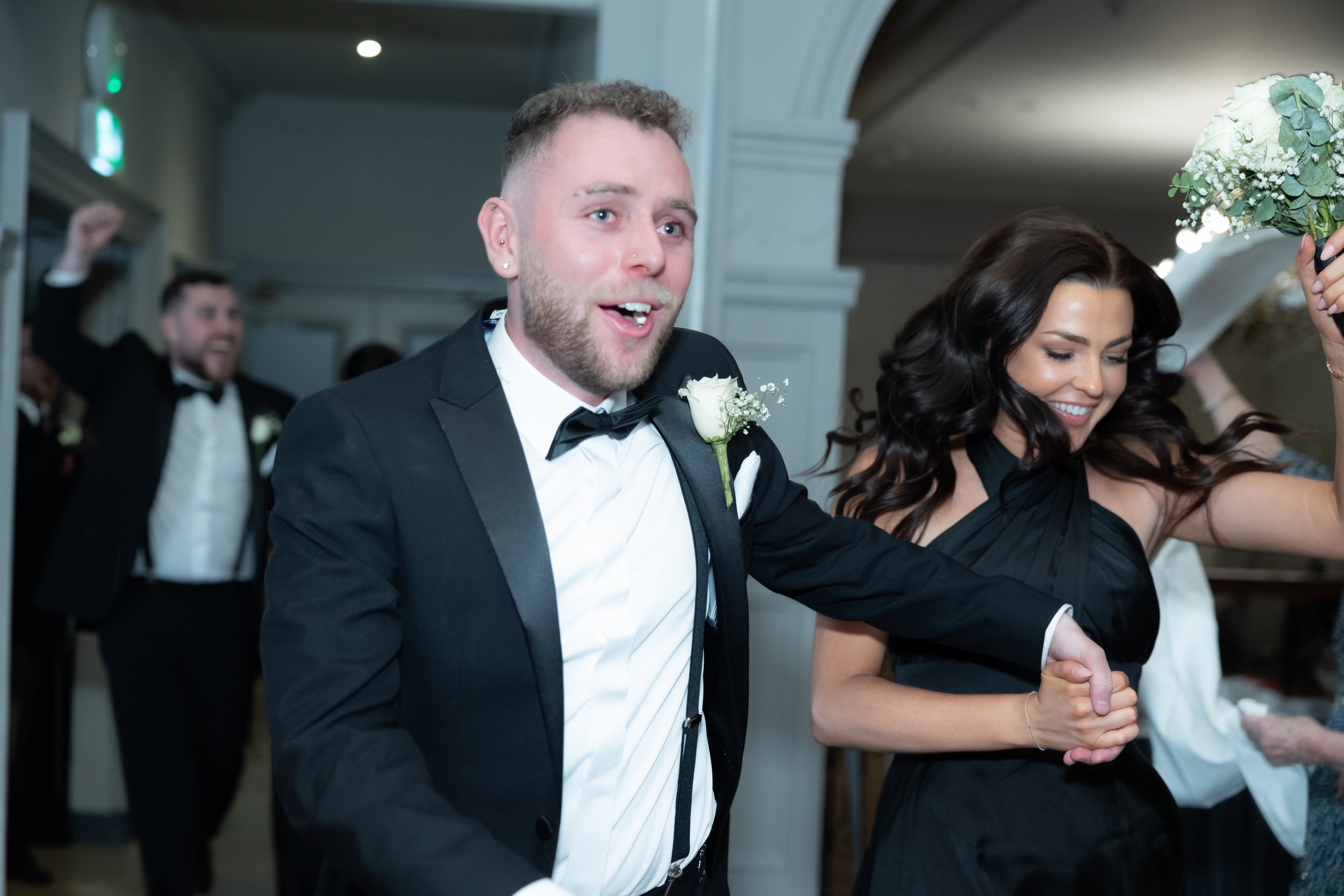A joyful couple dancing at a wedding reception, dressed in formal attire, with other guests in the background celebrating.