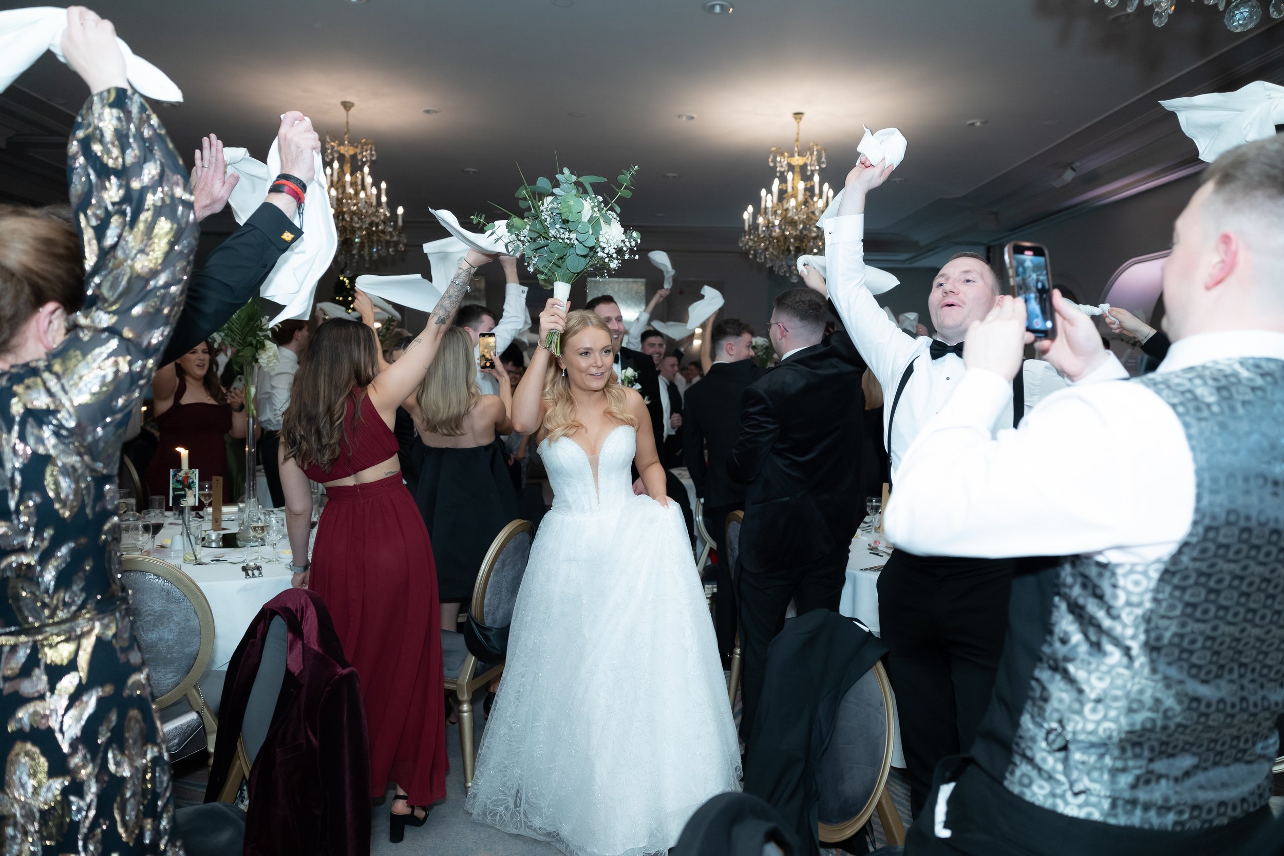 Bride in a white wedding gown holding a bouquet of flowers, surrounded by wedding guests celebrating and throwing napkins in the air at a reception hall with chandeliers.