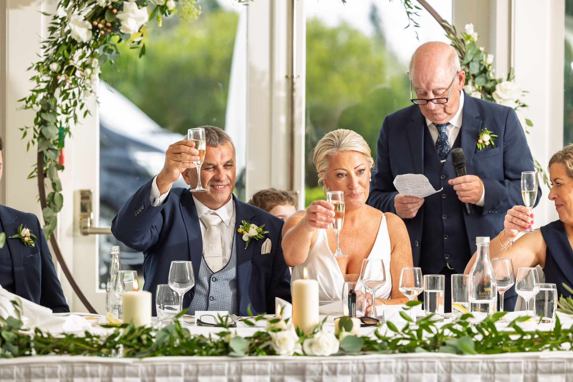 A wedding reception scene with guests raising glasses for a toast, seated at a decorated table with candles and flowers, and a man giving a speech using a microphone.