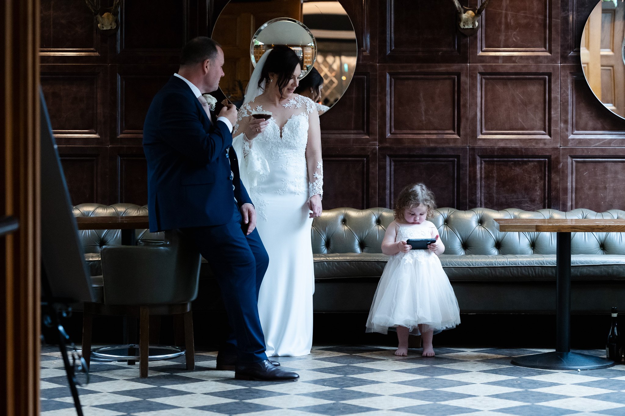 A bride and groom in wedding attire stand in a room with dark wood paneling, a tufted leather bench, and checkered floor tiles. The bride is looking at her phone, while the groom is drinking. A young girl in a white dress is standing barefoot, focuse