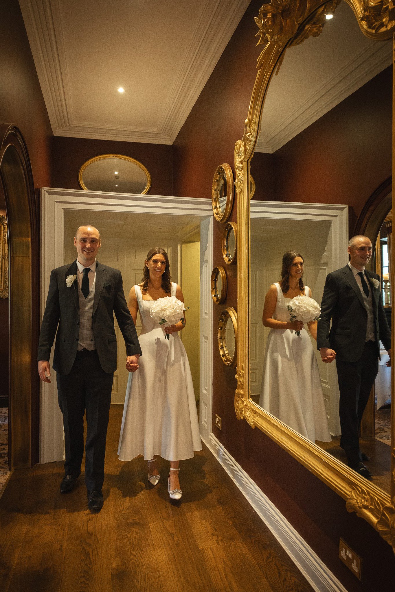 A bride and groom walking holding hands in a room with dark red walls, a large decorative mirror, and ornate gold-framed mirrors on the wall.