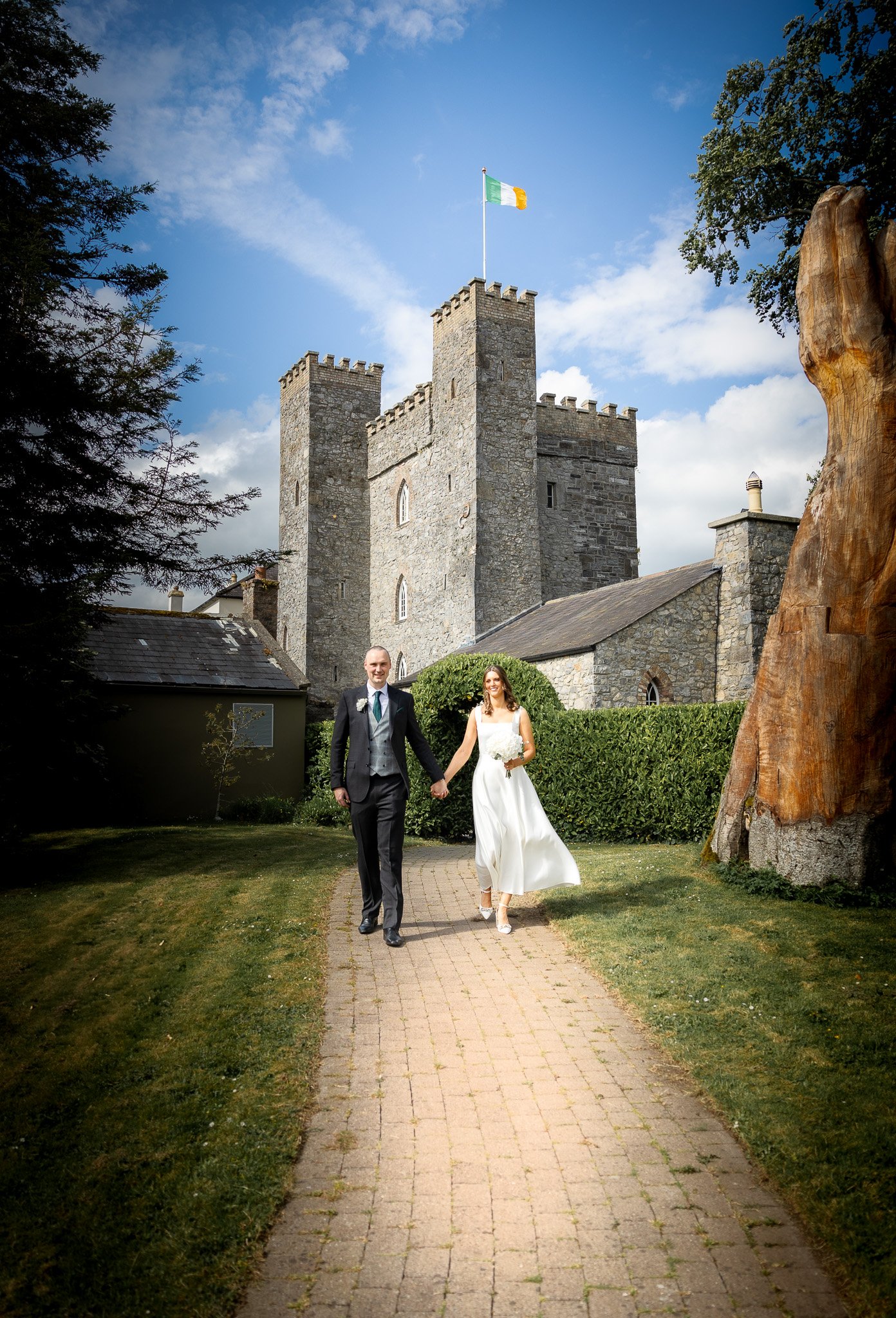 A bride and groom holding hands and walking on a brick path in front of a historic stone castle under a partly cloudy sky.