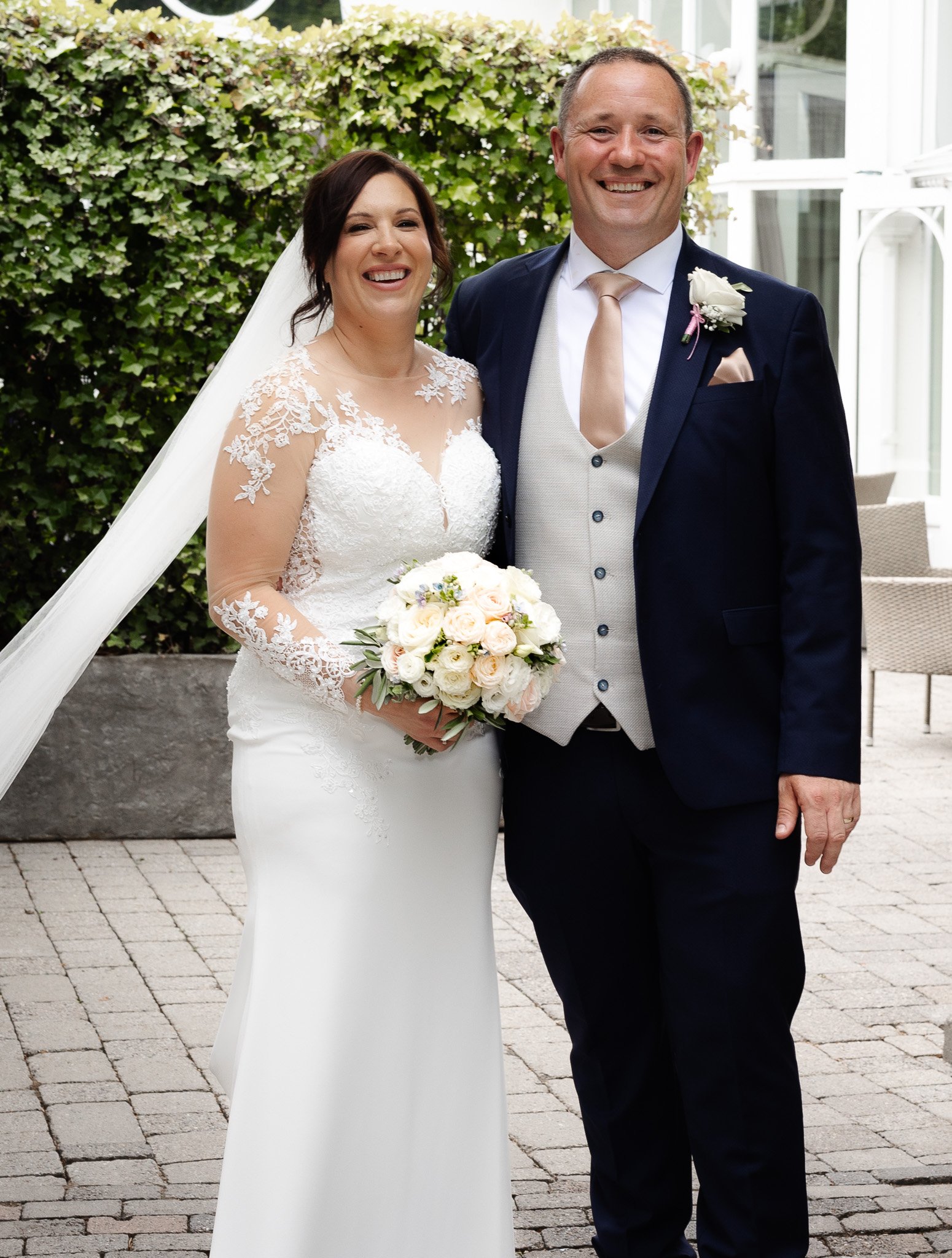 Happy bride and groom standing outside, smiling, with the bride holding a bouquet of roses, in wedding attire, near a green leafy hedge and white conservatory-like building.