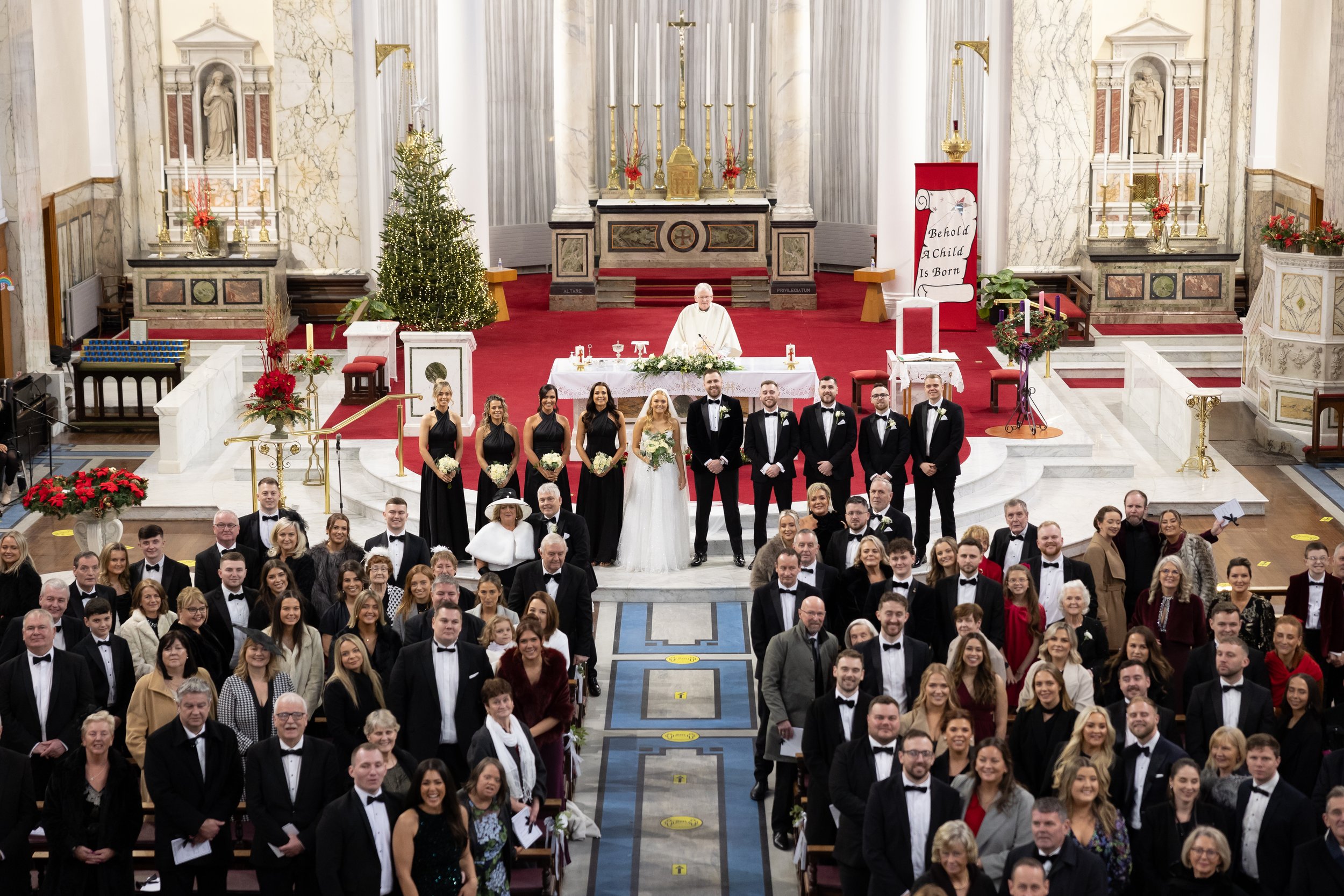A large wedding ceremony inside a church with a bride and groom at the center, surrounded by bridesmaids and groomsmen, with guests seated in the pews.