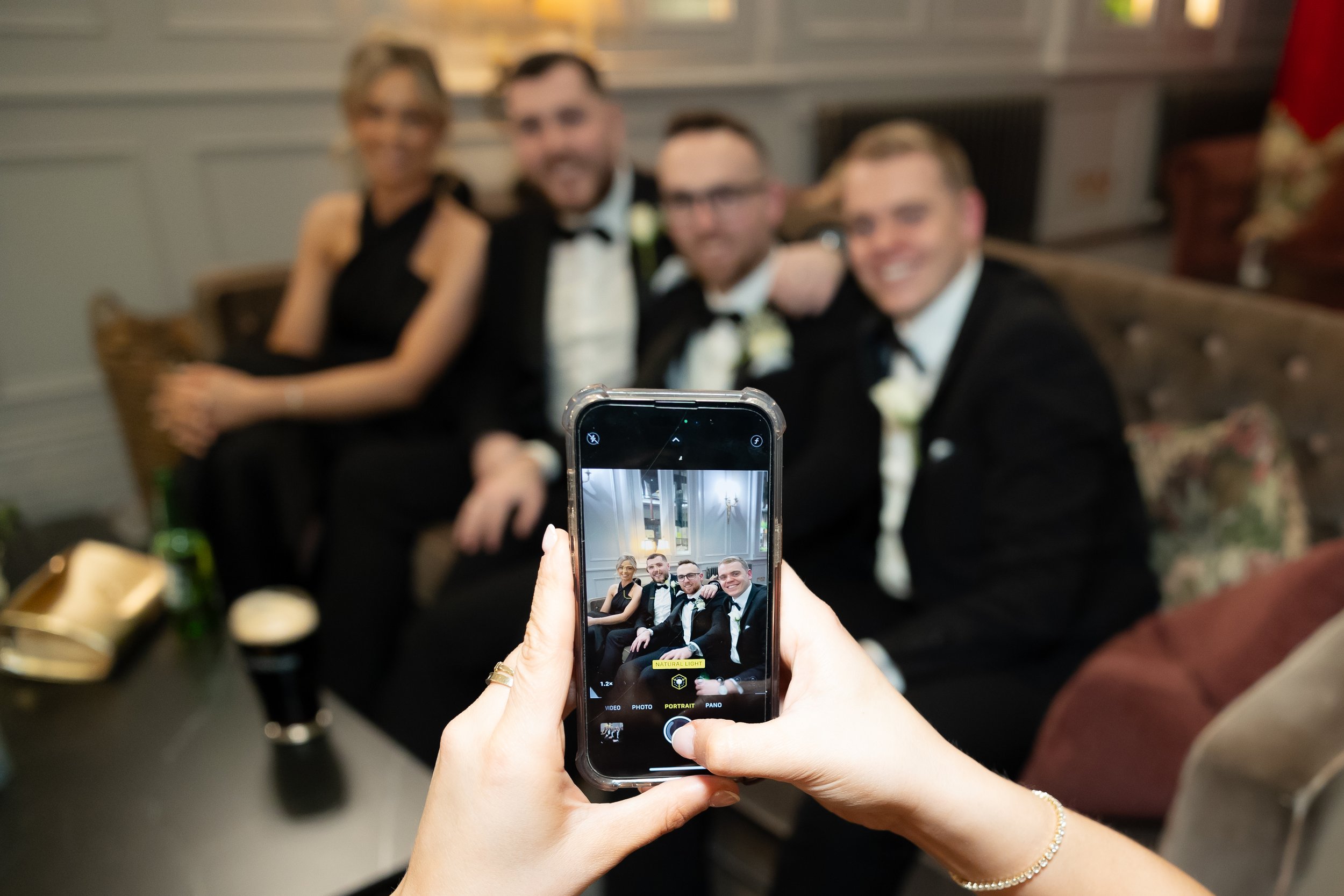 Person taking a photo of four people dressed in formal wear on a couch with a smartphone.