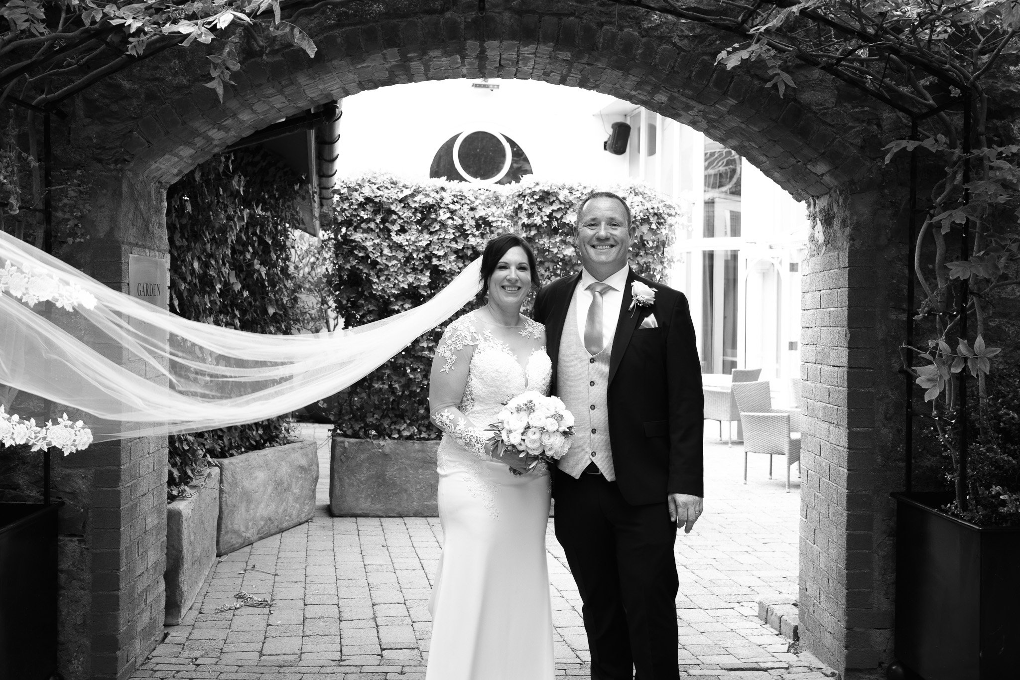 A bride and groom standing together outside under a brick archway, smiling, with the bride holding a bouquet of flowers.