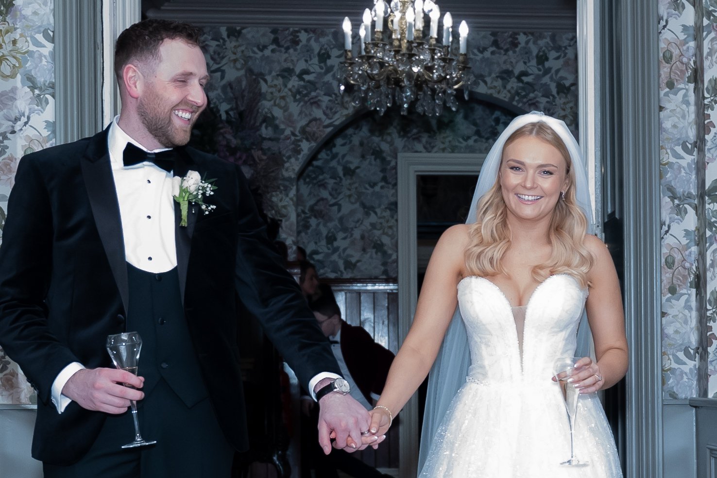 A bride and groom in wedding attire holding hands and smiling at each other inside a decorated room with a chandelier and floral wallpaper.