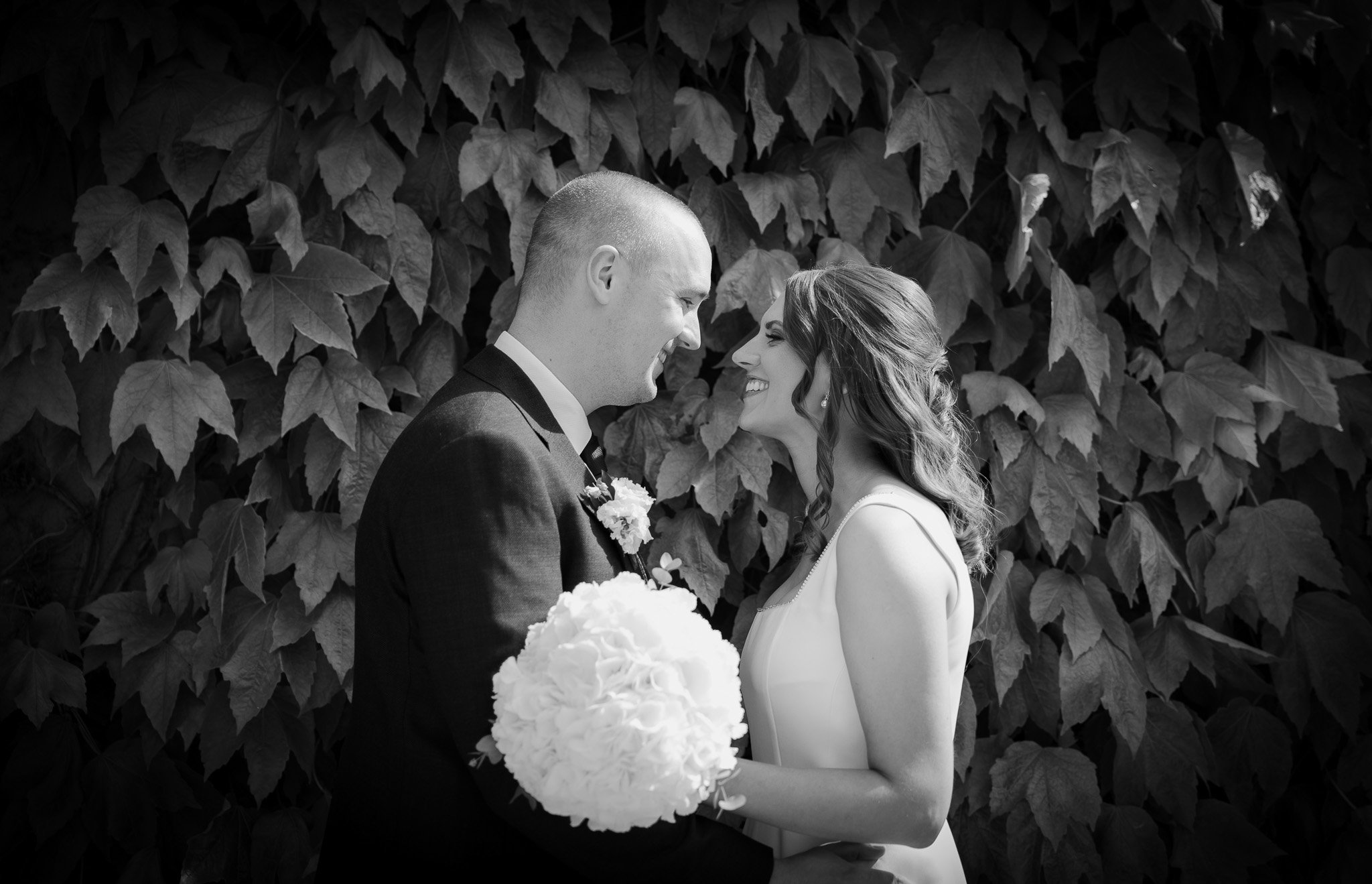 Black and white photo of a bride and groom smiling and facing each other outdoors, standing in front of a background of large leafy plants.