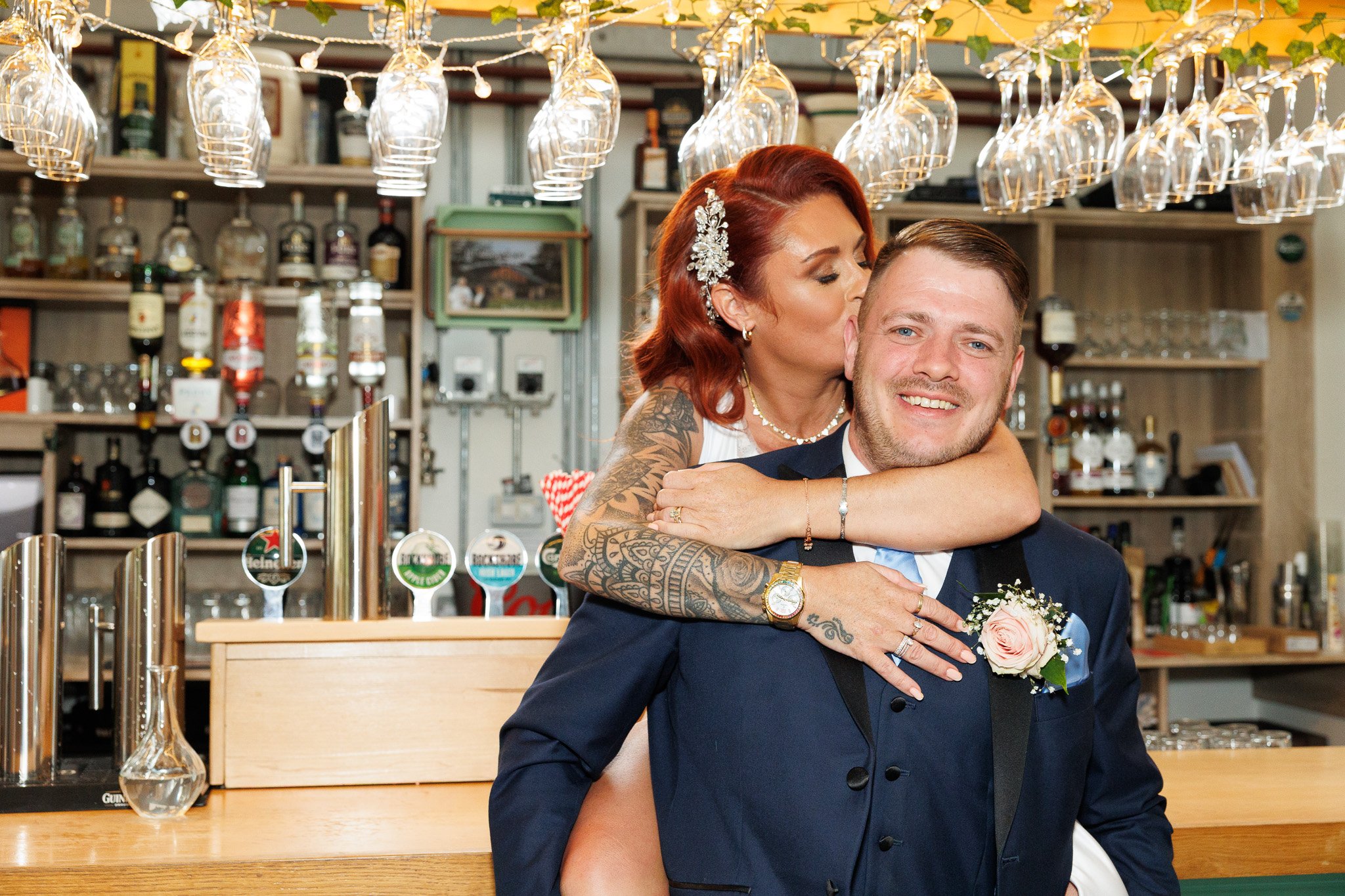 A woman with red hair and tattoos is giving a kiss on the cheek to a smiling man in a suit, at a bar setting with hanging wine glasses and bottles in the background.