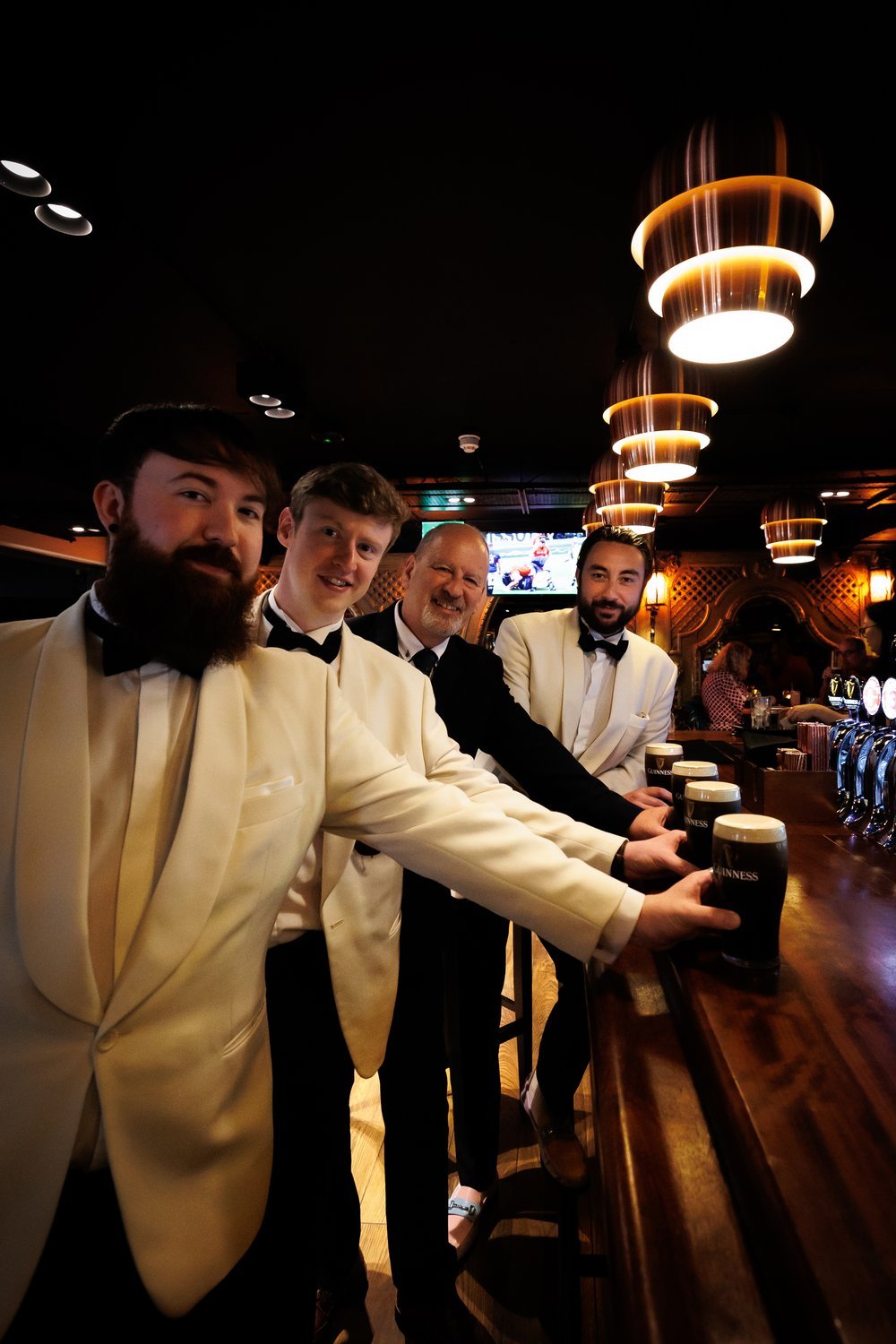 Four men dressed in formal tuxedos, three in cream-colored jackets and one in a black jacket, are standing at a bar in a dimly lit pub, holding glasses of Guinness beer. The background features warm lighting, a television screen, and other patrons.