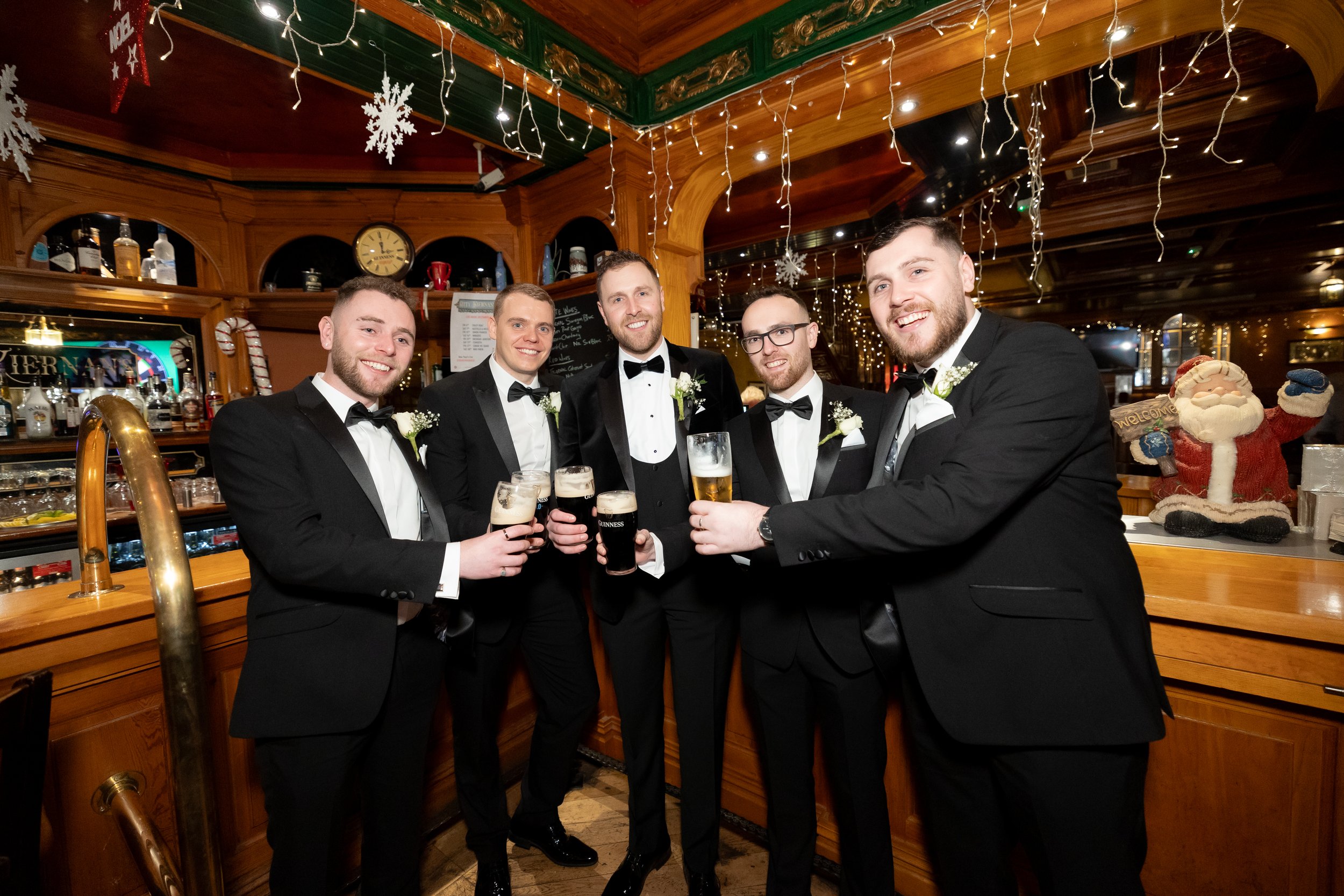 Group of six men in tuxedos holding drinks, smiling at a bar decorated with Christmas lights and ornaments, including a Santa Claus figure in the background.