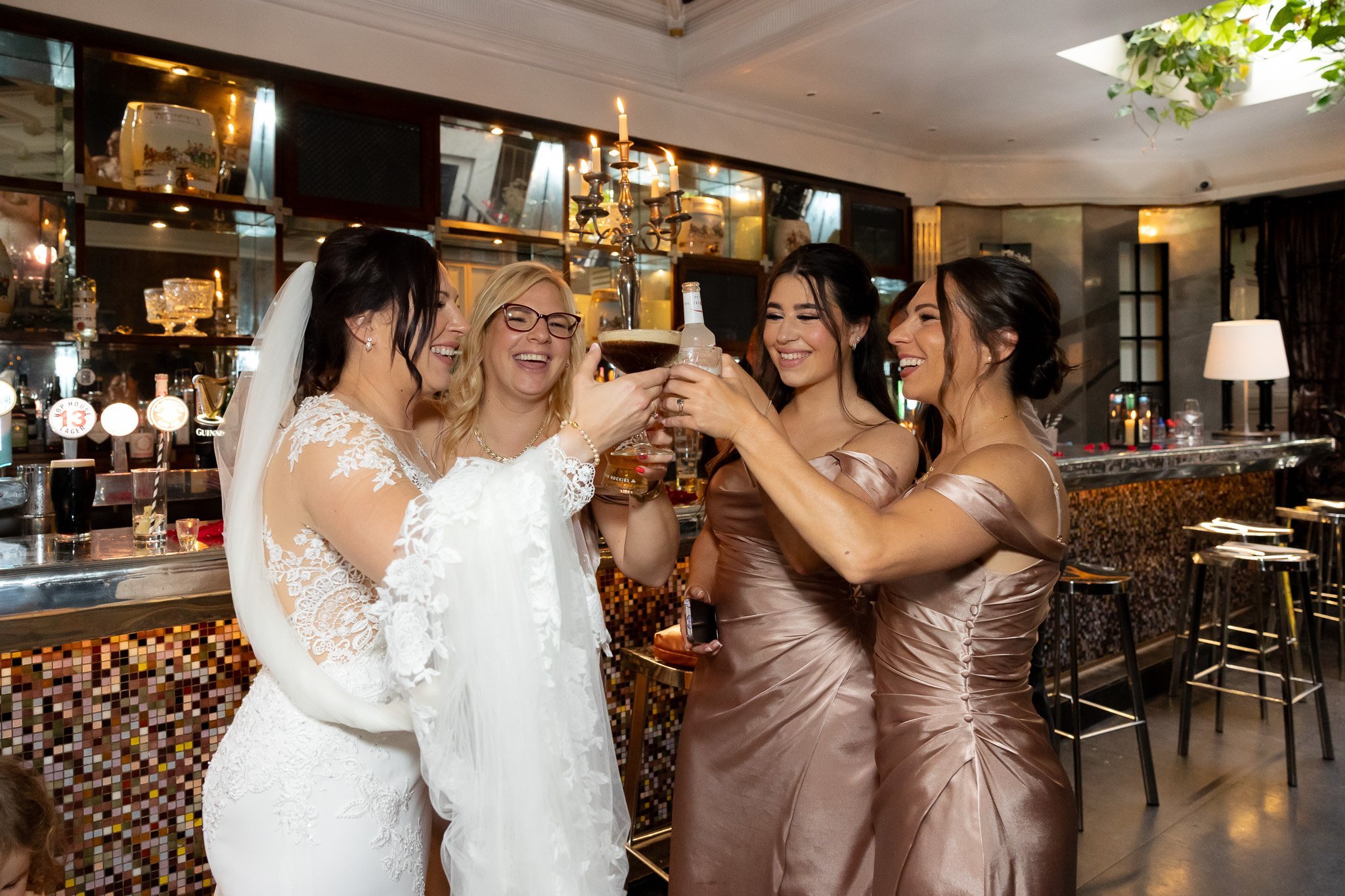 Group of women celebrating at a bar, raising drinks and smiling, including a bride in white wedding dress and veil, with three bridesmaids in satin dresses.