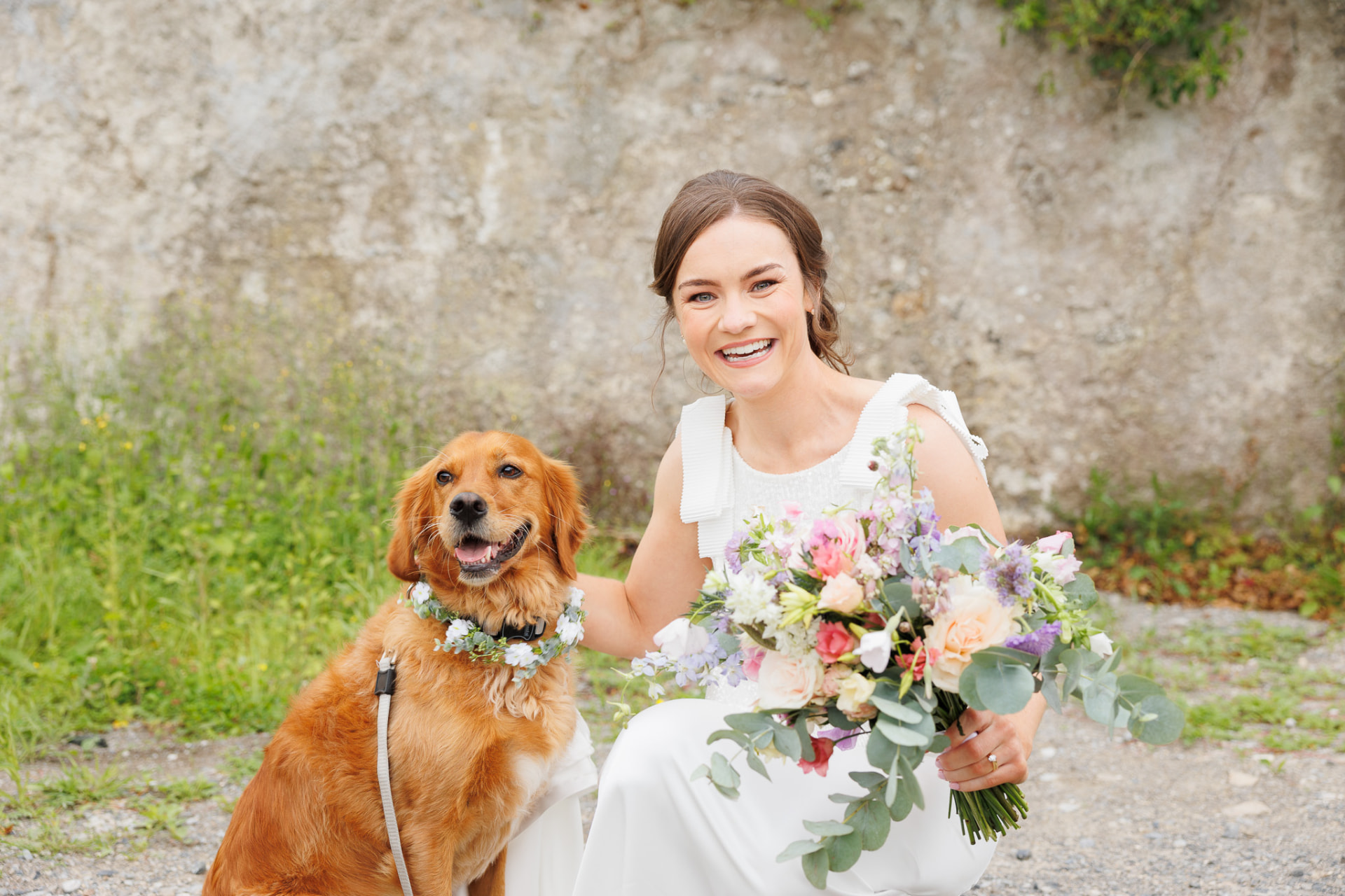 Bride kneeling beside her golden retriever with a wildflower bouquet