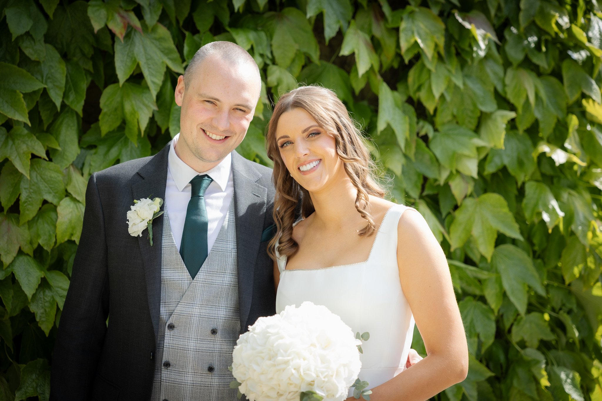 A smiling bride and groom standing outdoors in front of green leaves, with the bride holding a white bouquet.
