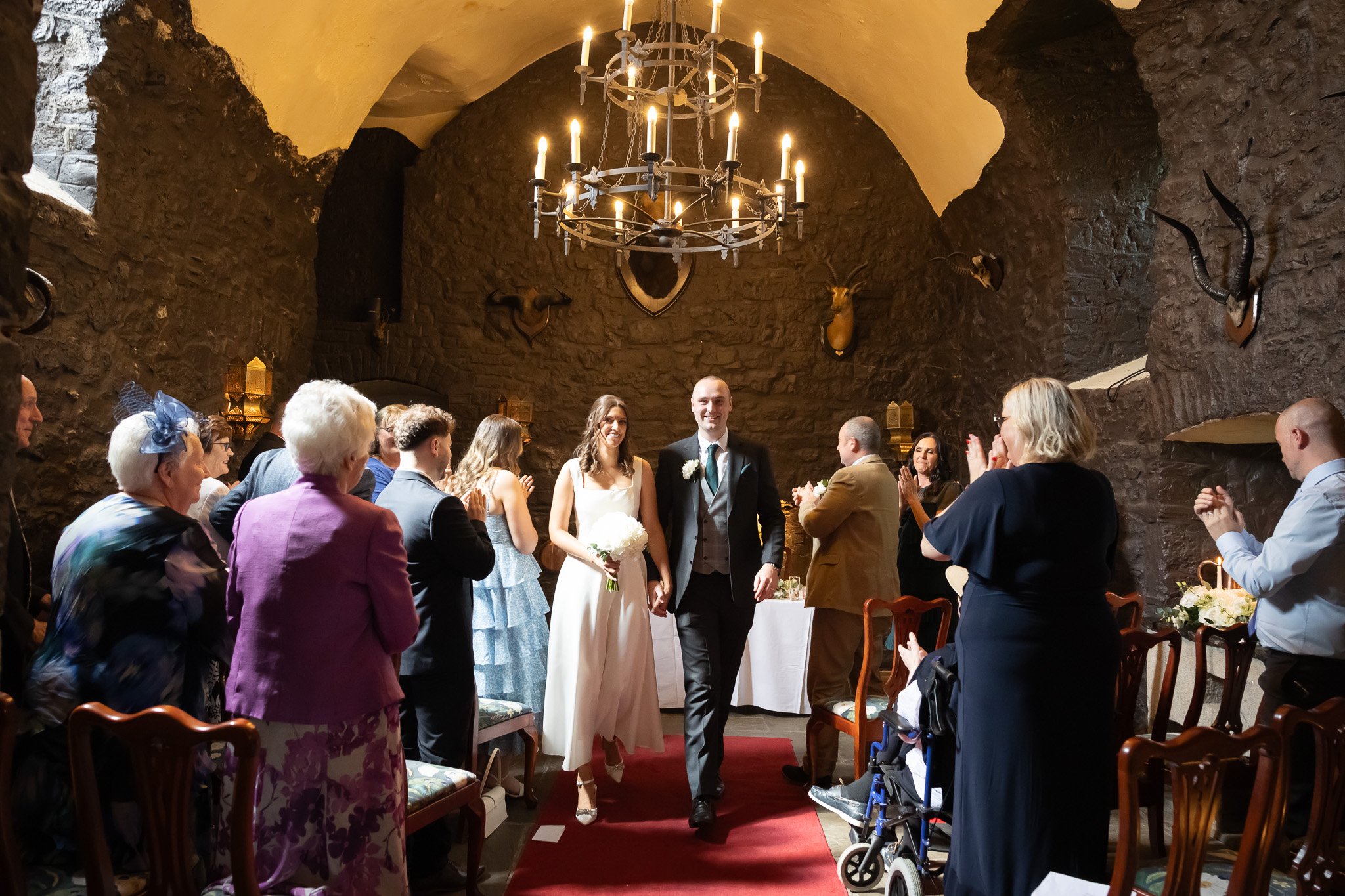 A wedding ceremony taking place in a stone room with a chandelier and mounted animal heads on the walls. A bride and groom are walking down the aisle while guests on both sides applaud.