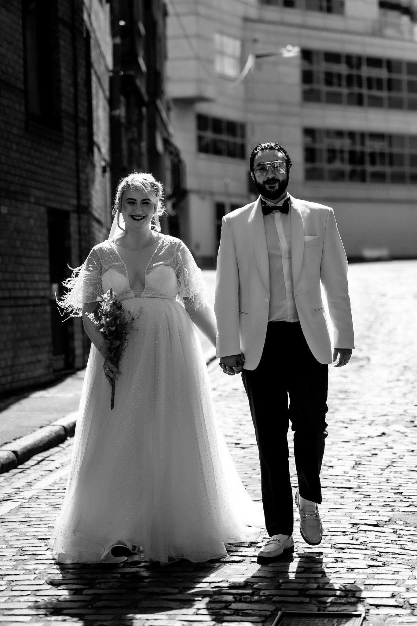 Black and white photo of a bride and groom walking hand in hand on a cobblestone street, smiling, with modern buildings in the background.