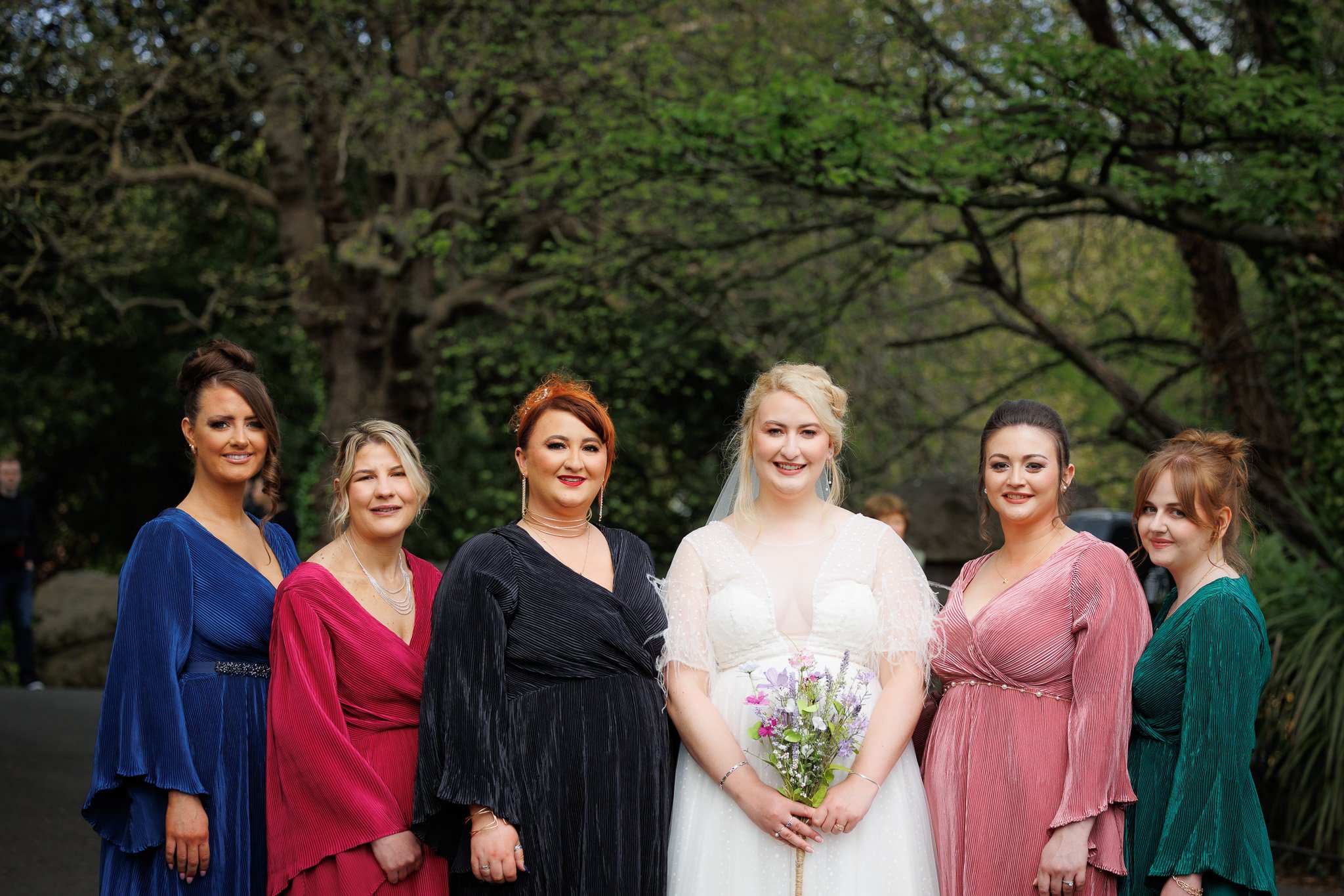 Group of six women, including a bride in a white gown holding a bouquet, standing outdoors with green trees in the background.