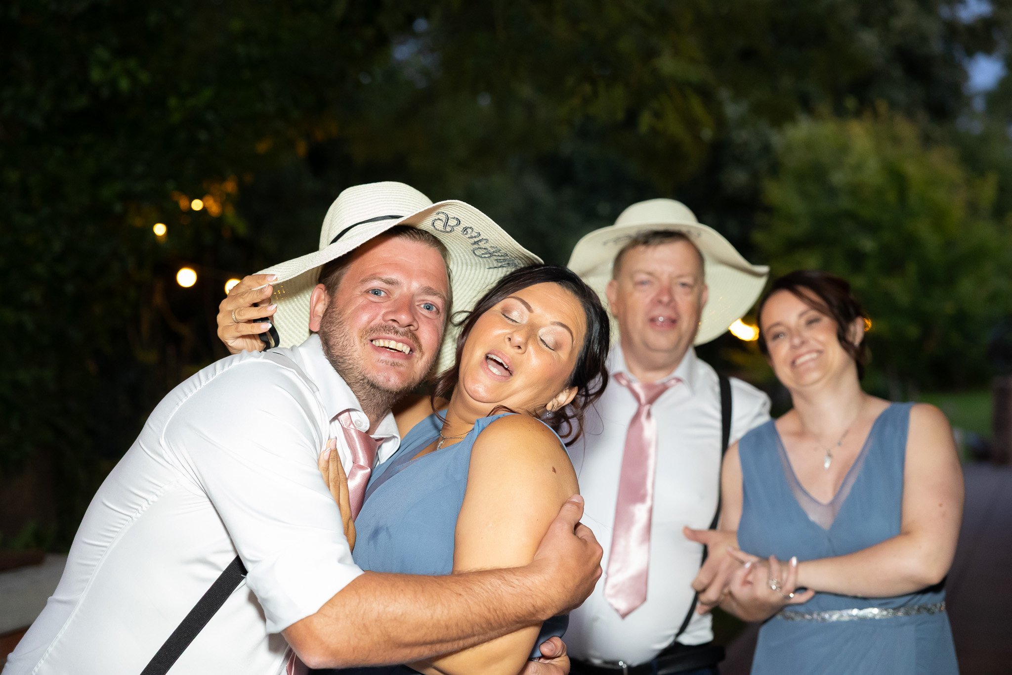 Group of four adults celebrating at an outdoor event during evening, wearing hats and semi-formal attire, with trees and lights in the background.