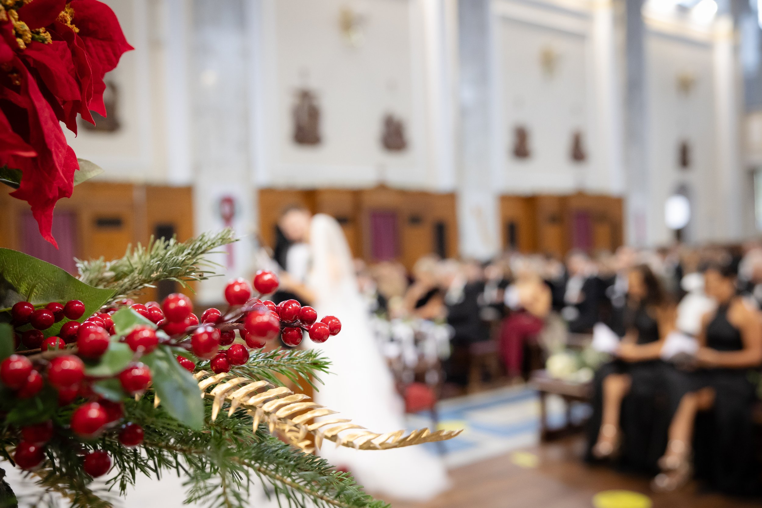 Close-up of Christmas decorations with red berries and greenery, blurred background showing a church interior filled with attendees, a bride and groom at the altar.