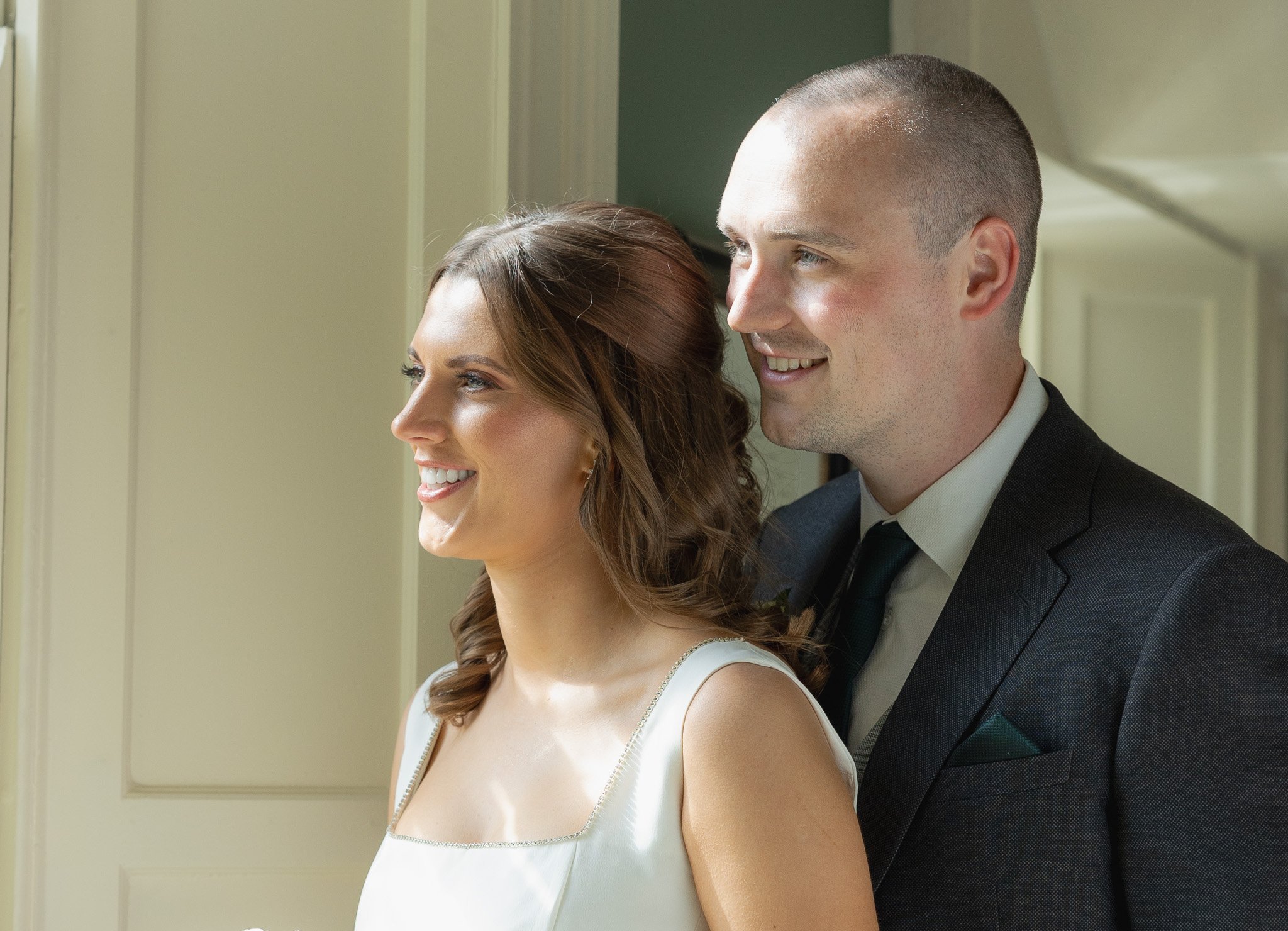 A bride and groom standing together indoors, smiling and looking out a window.