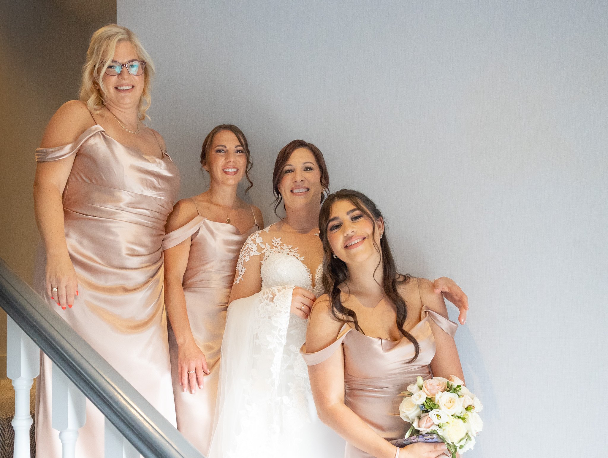 A bride and four bridesmaids posing together on a staircase, with the bride holding a bouquet of white and pink roses. All are smiling and wearing matching blush-colored dresses, with a plain white wall as background.