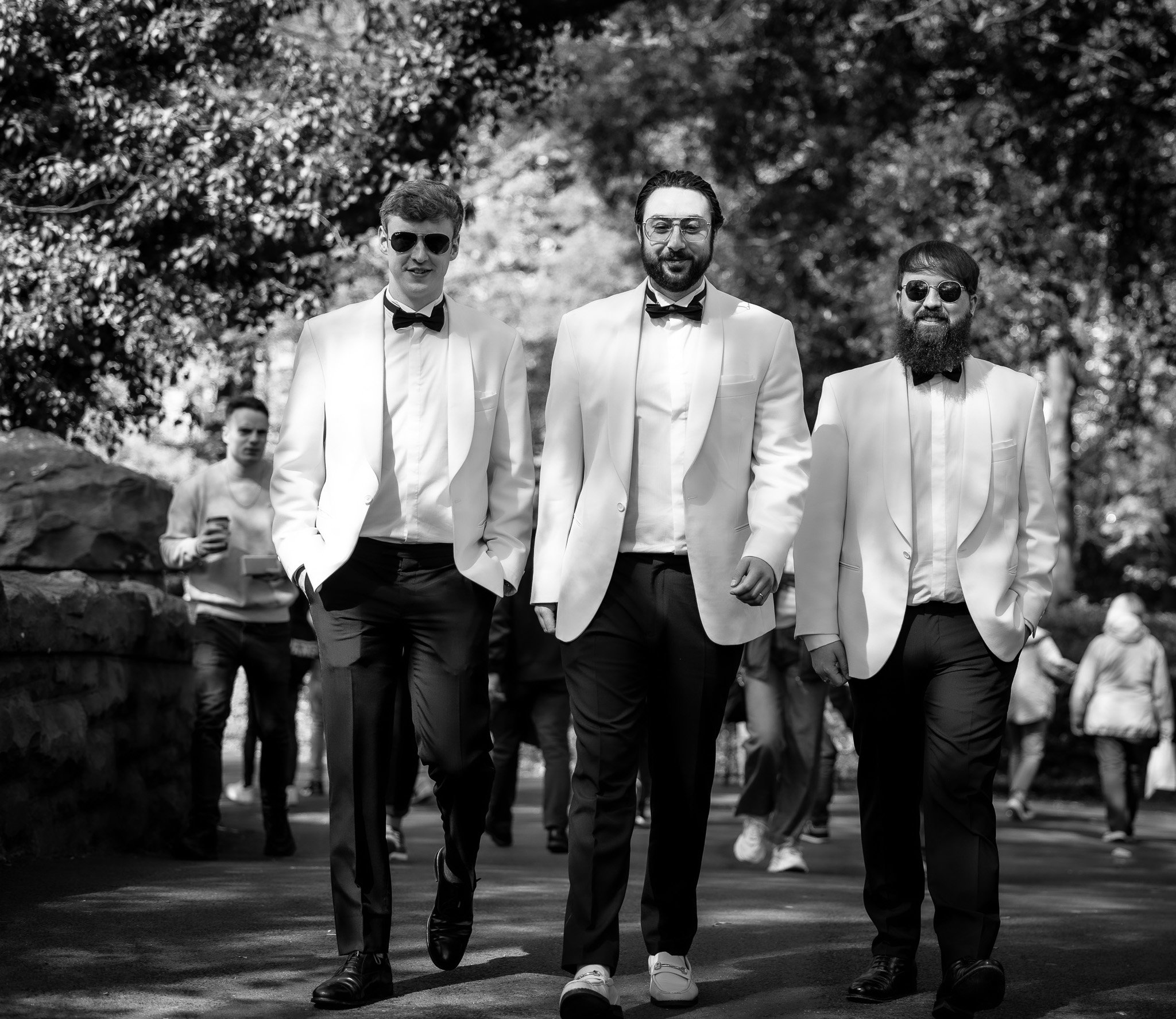 Three men in white tuxedos and black bow ties walking outdoors, with trees and other people in the background.