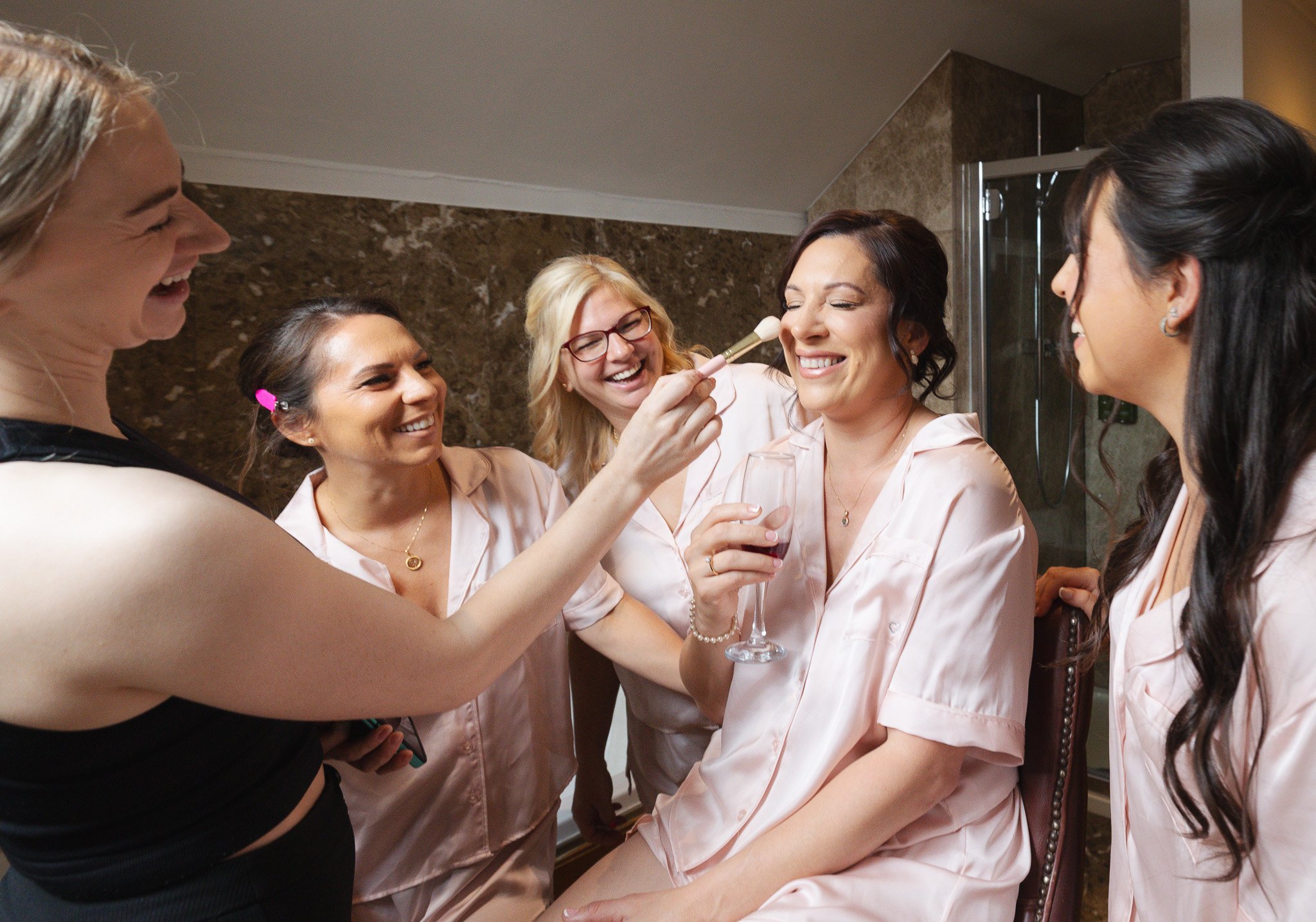 A bride gets her makeup done by friends during a bridal party, with one friend applying blush and others smiling and celebrating.