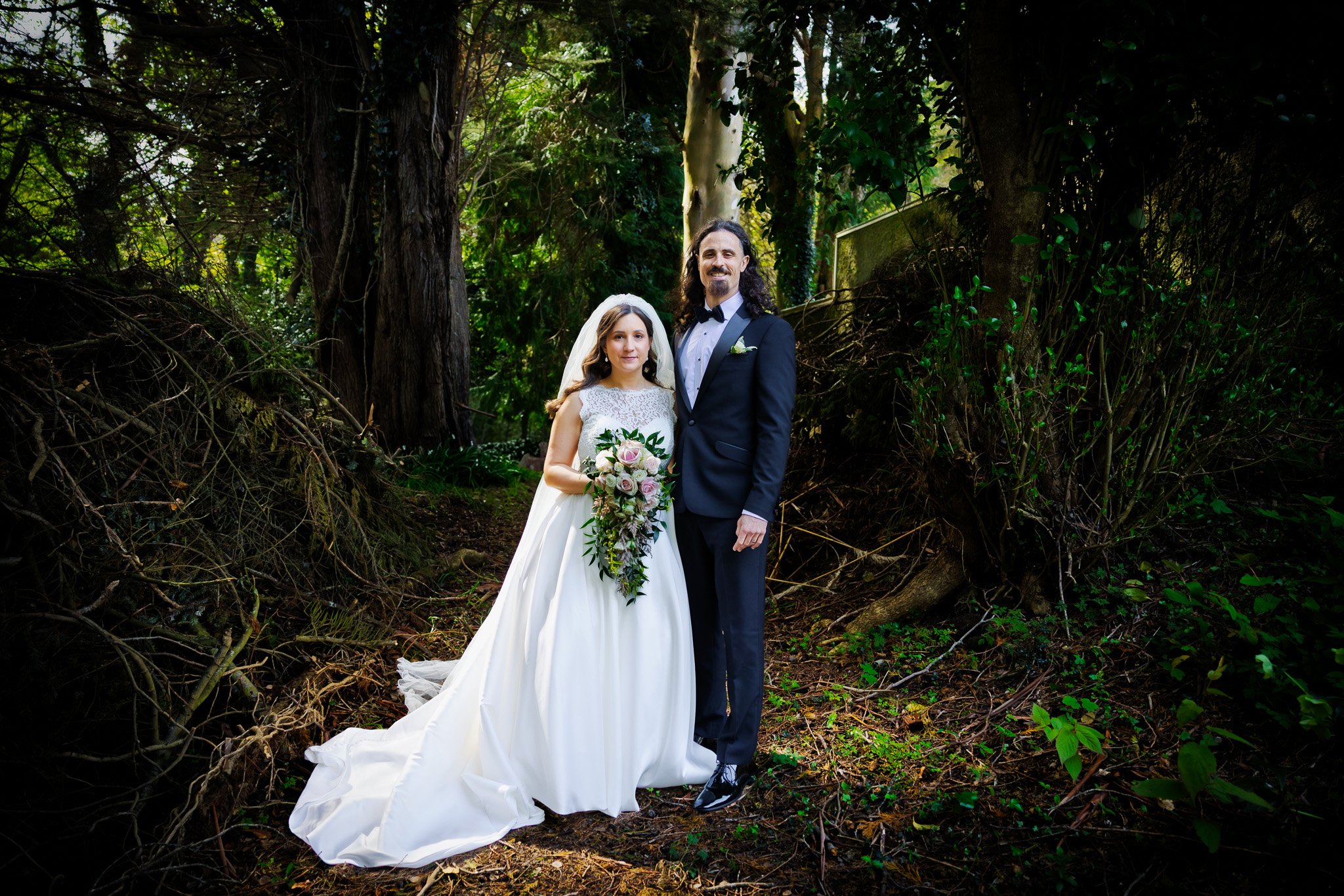 A bride and groom dressed in wedding attire standing in a forest, with trees and green foliage surrounding them.